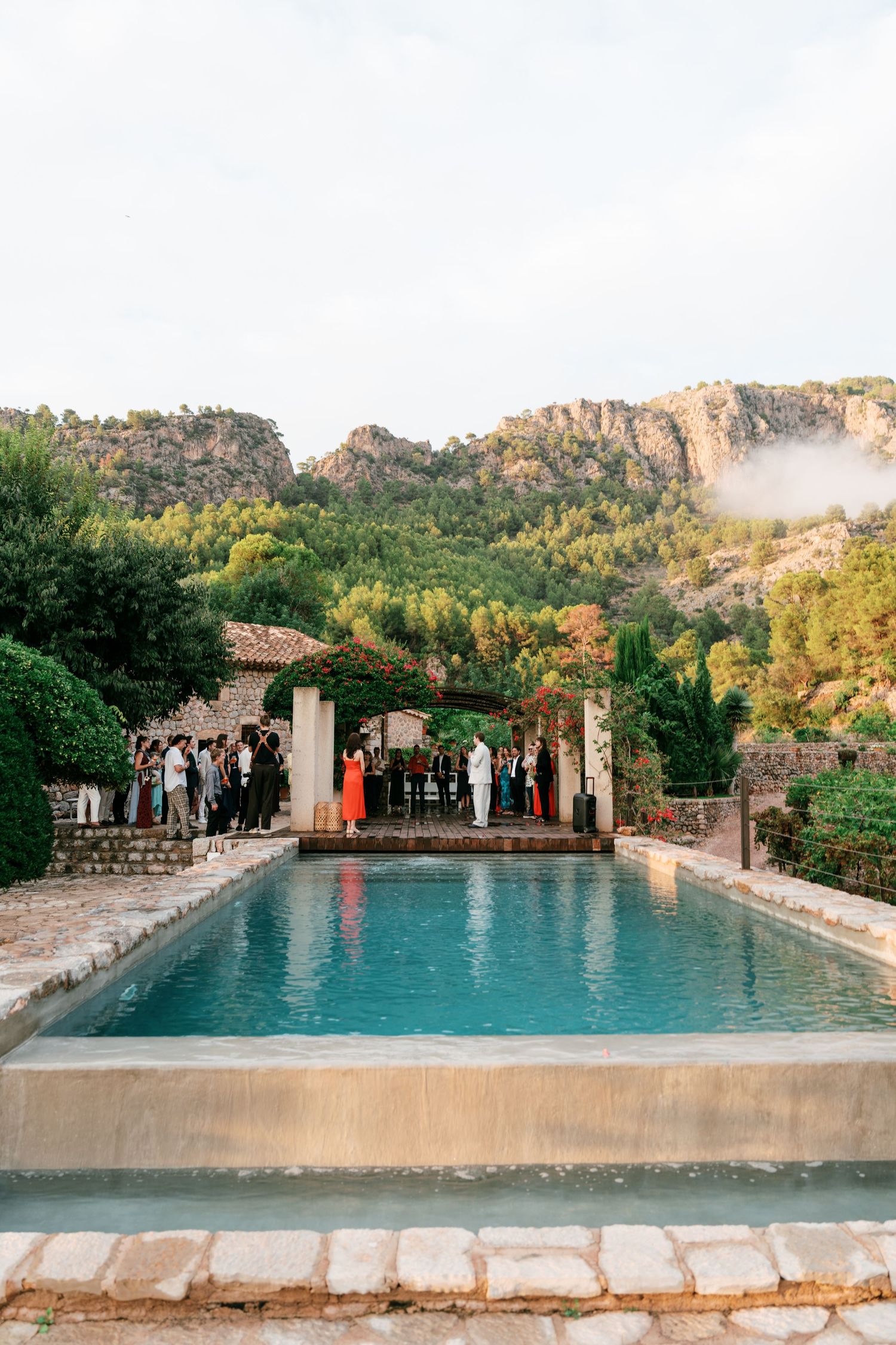 Wedding guests gather under a vine-covered pergola beside a turquoise pool, with rugged green mountains rising in the background.