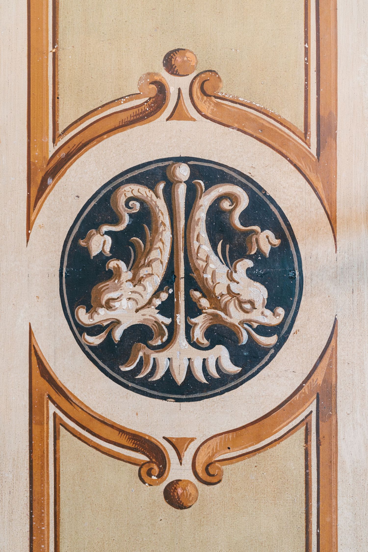 Decorative painted panel with two symmetrical dolphins flanking a central trident in a dark circular medallion, framed by ornate scrollwork.
