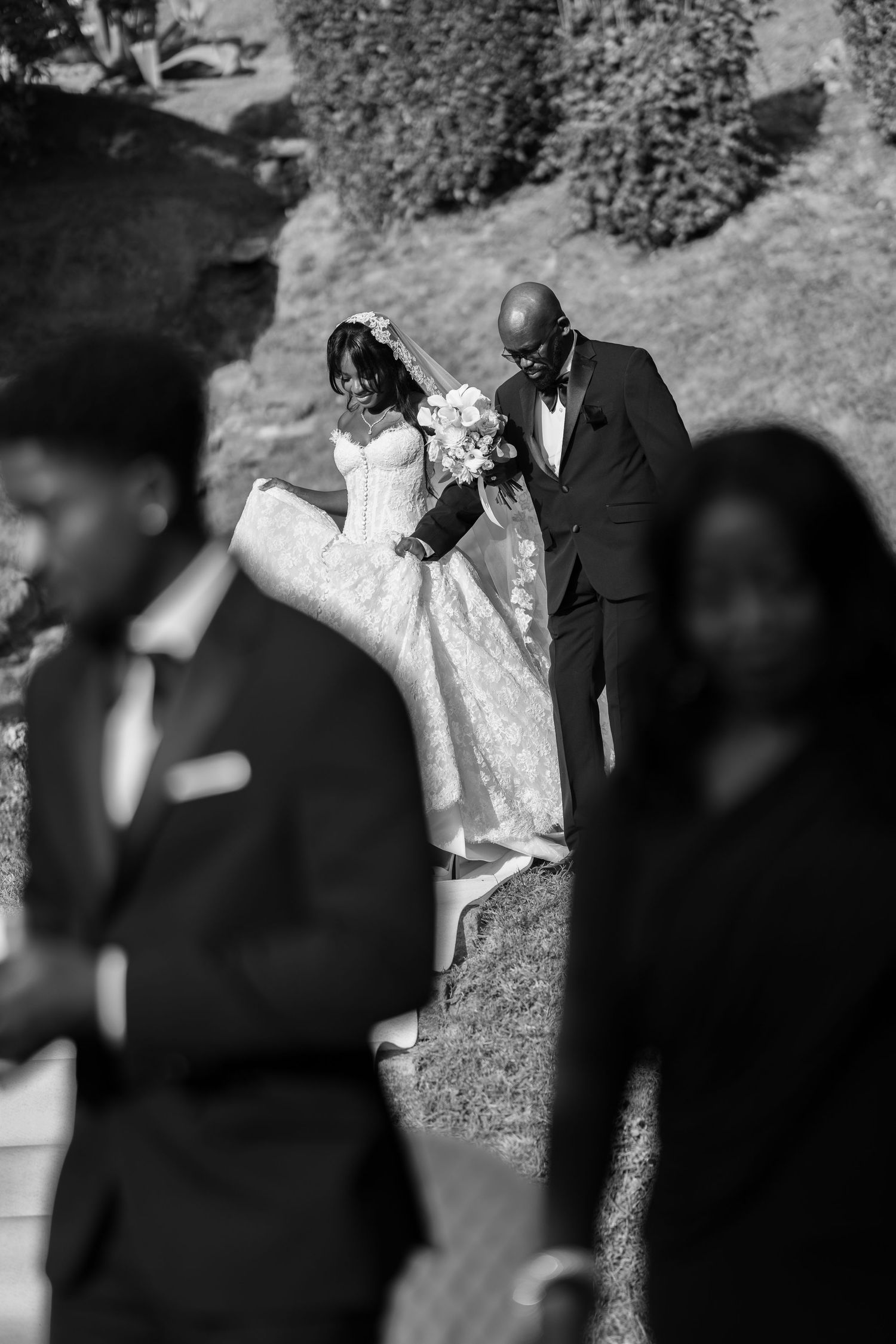 Black and white photo of a bride in a lace gown holding a bouquet, walking down a grassy aisle with a man in a tuxedo at a wedding.
