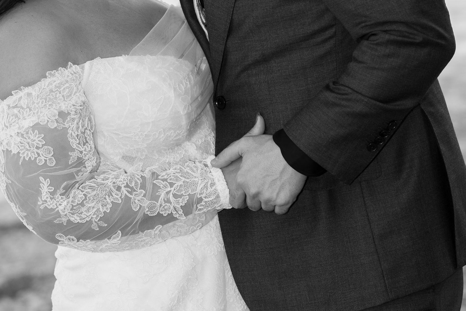 Black and white close-up of a bride in a lace long-sleeve wedding gown embracing a groom in a suit, his hand resting gently on her waist.