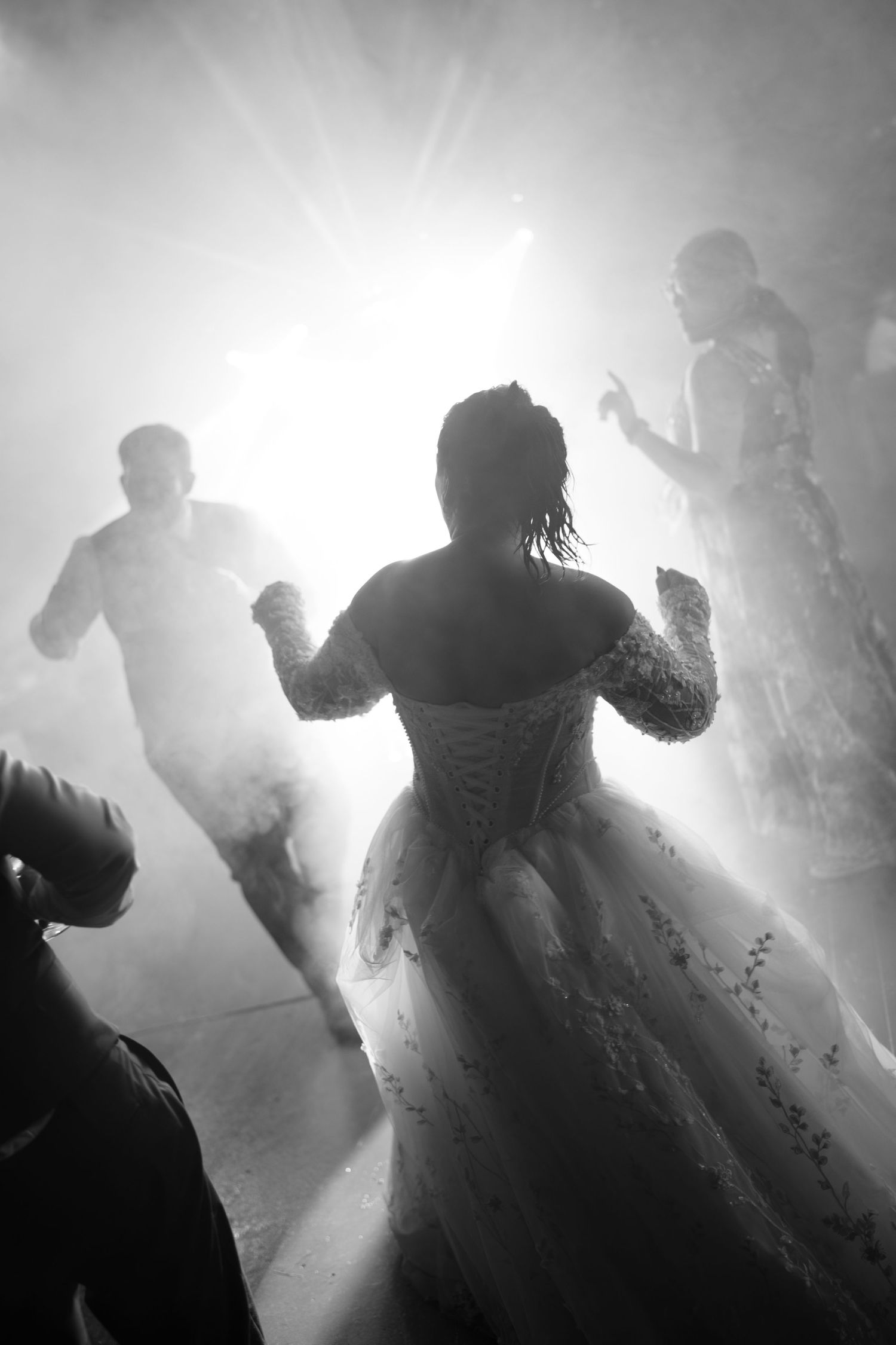 Black and white photo of a bride in a lace-up corset gown dancing with guests amid bright backlight and haze.