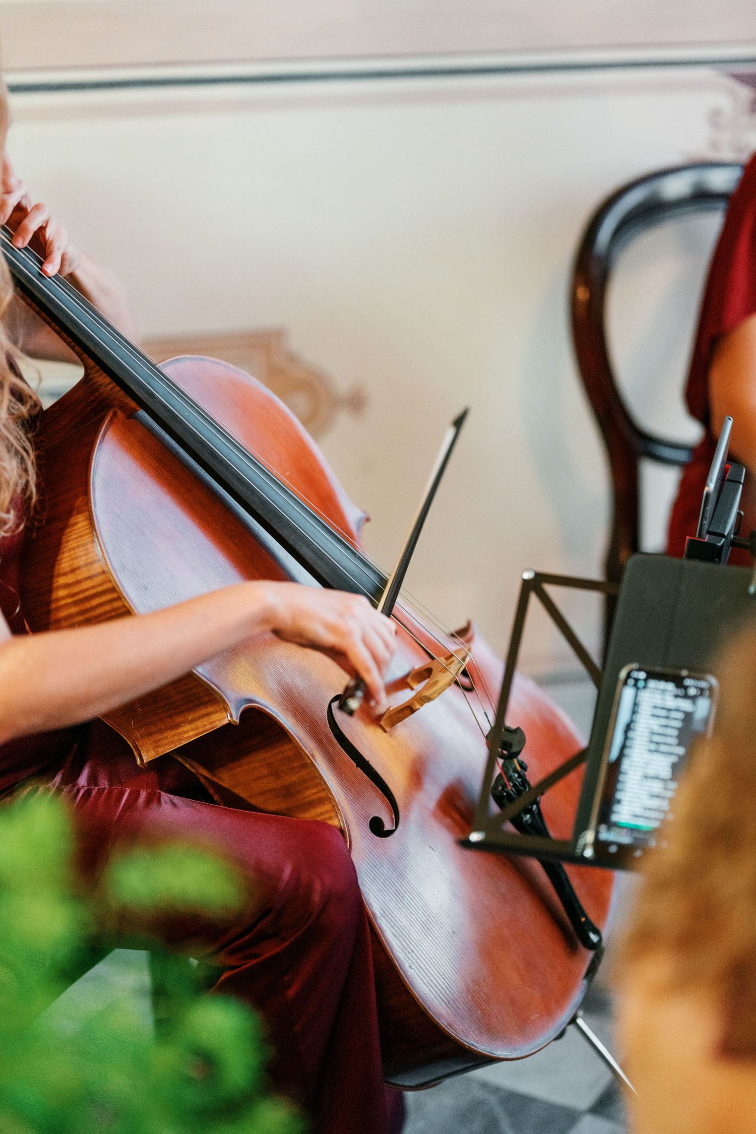 Cellist in a burgundy outfit holds a bow against a wooden cello while sheet music sits on a stand during an indoor performance.