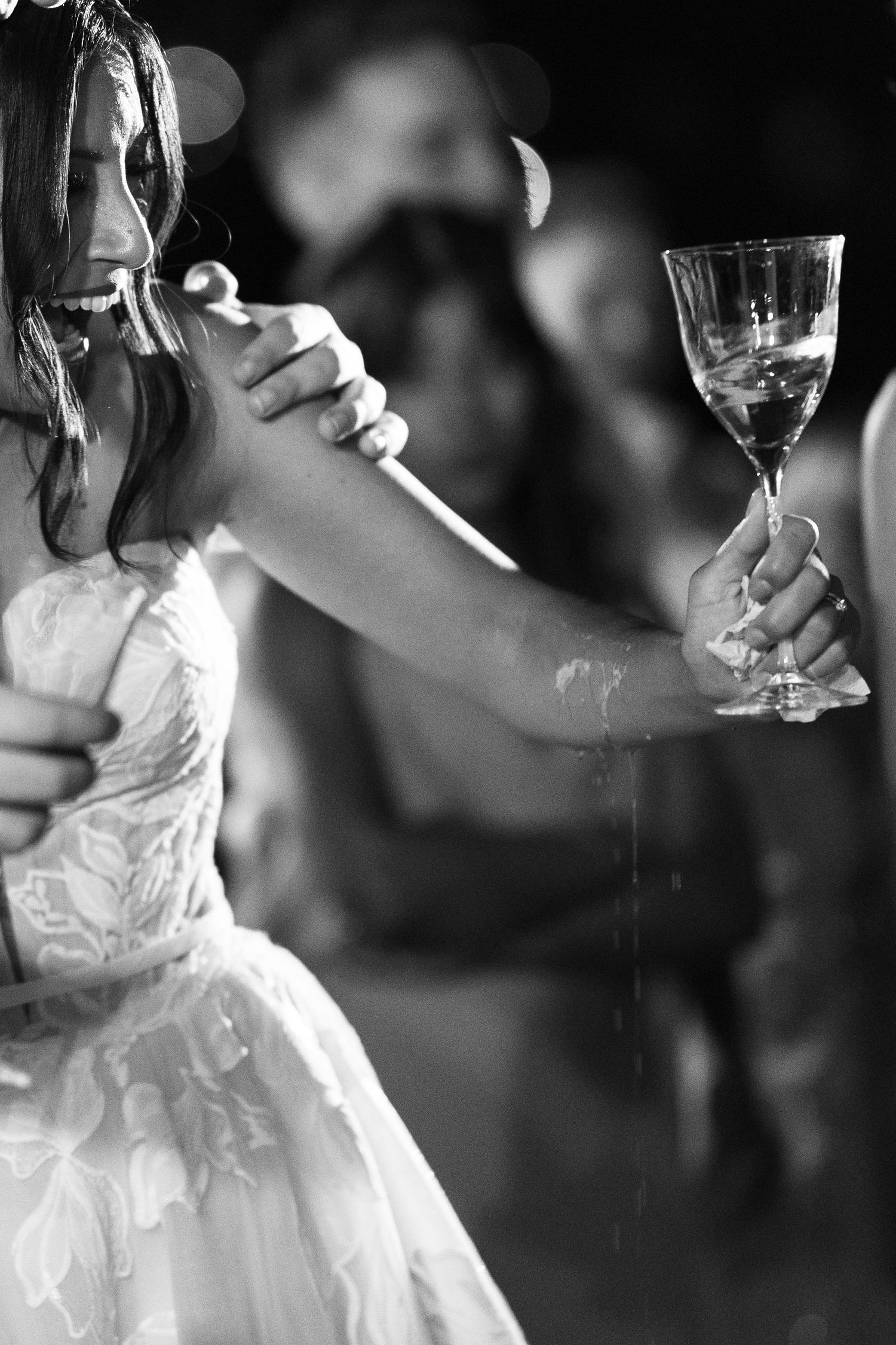Laughing bride in a lace wedding gown raises a champagne glass as drops spill, with guests blurred behind her in a black and white photo.
