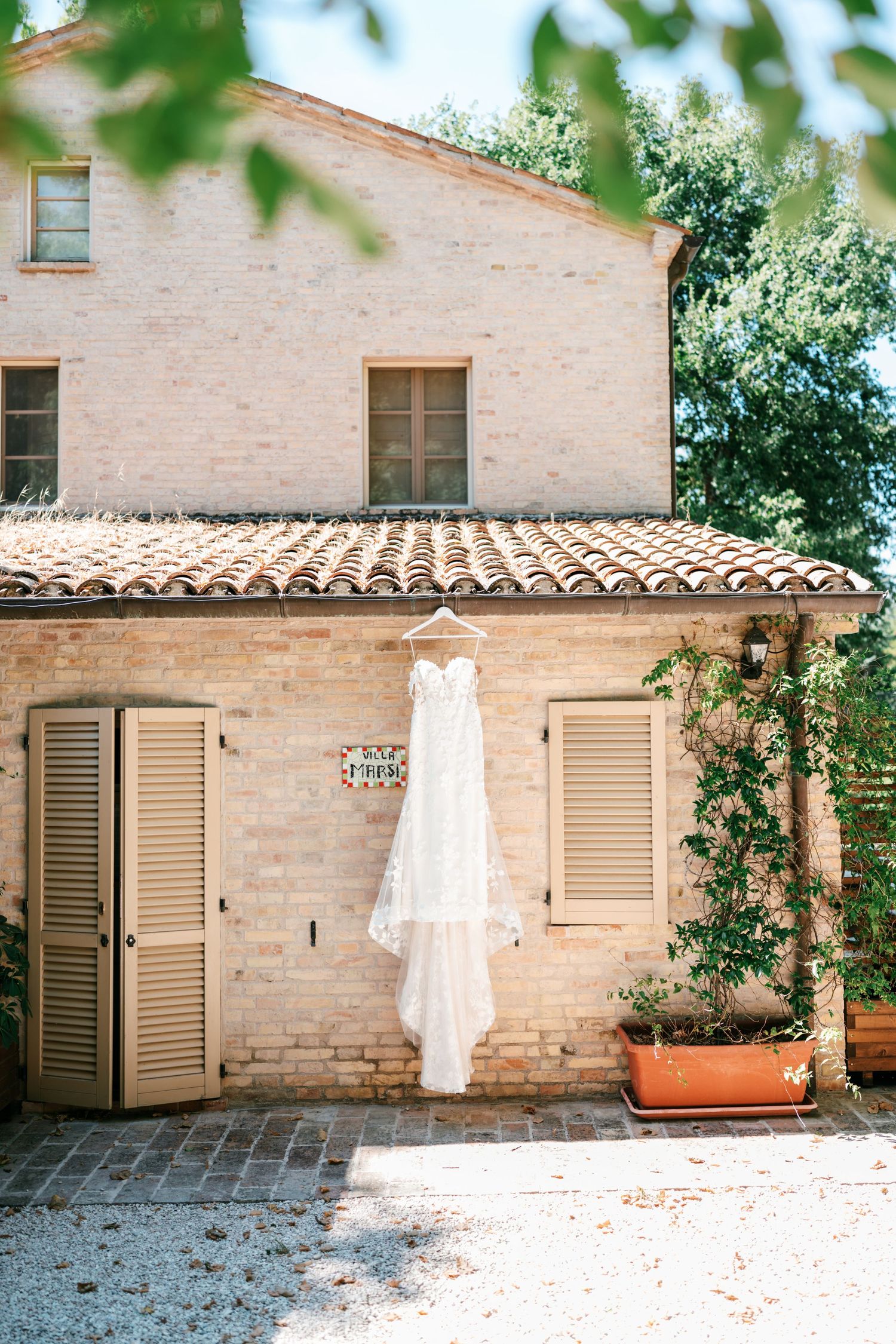 White wedding dress hanging on a rustic Italian villa facade with terracotta roof tiles and wooden shutters.