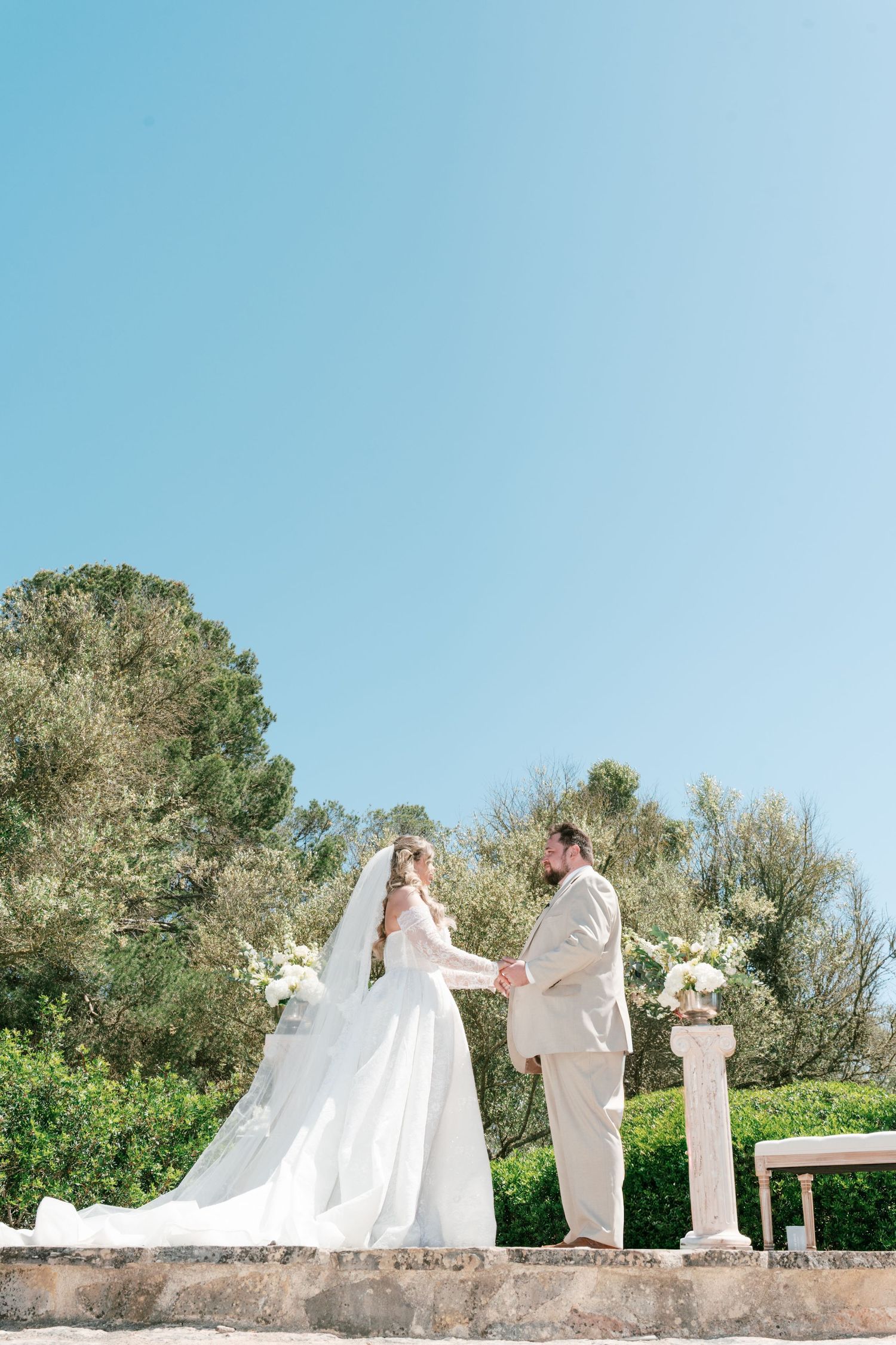 Bride and groom exchange vows outdoors at a sunlit wedding ceremony with white floral arrangements and lush greenery.