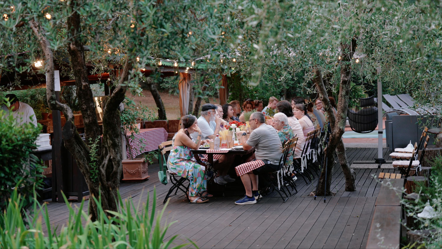 Group of friends gathers around a long wooden table on an outdoor deck beneath olive trees, enjoying a relaxed dinner at dusk.