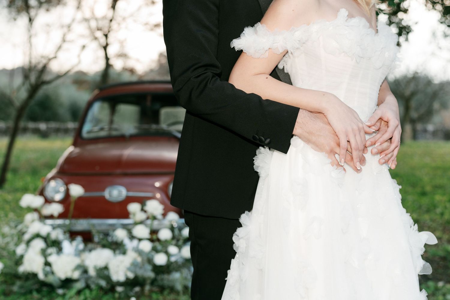 Bride and groom embracing near a vintage red Fiat 500 surrounded by white flowers in a green field. Mallorca wedding photographer, filmmaker