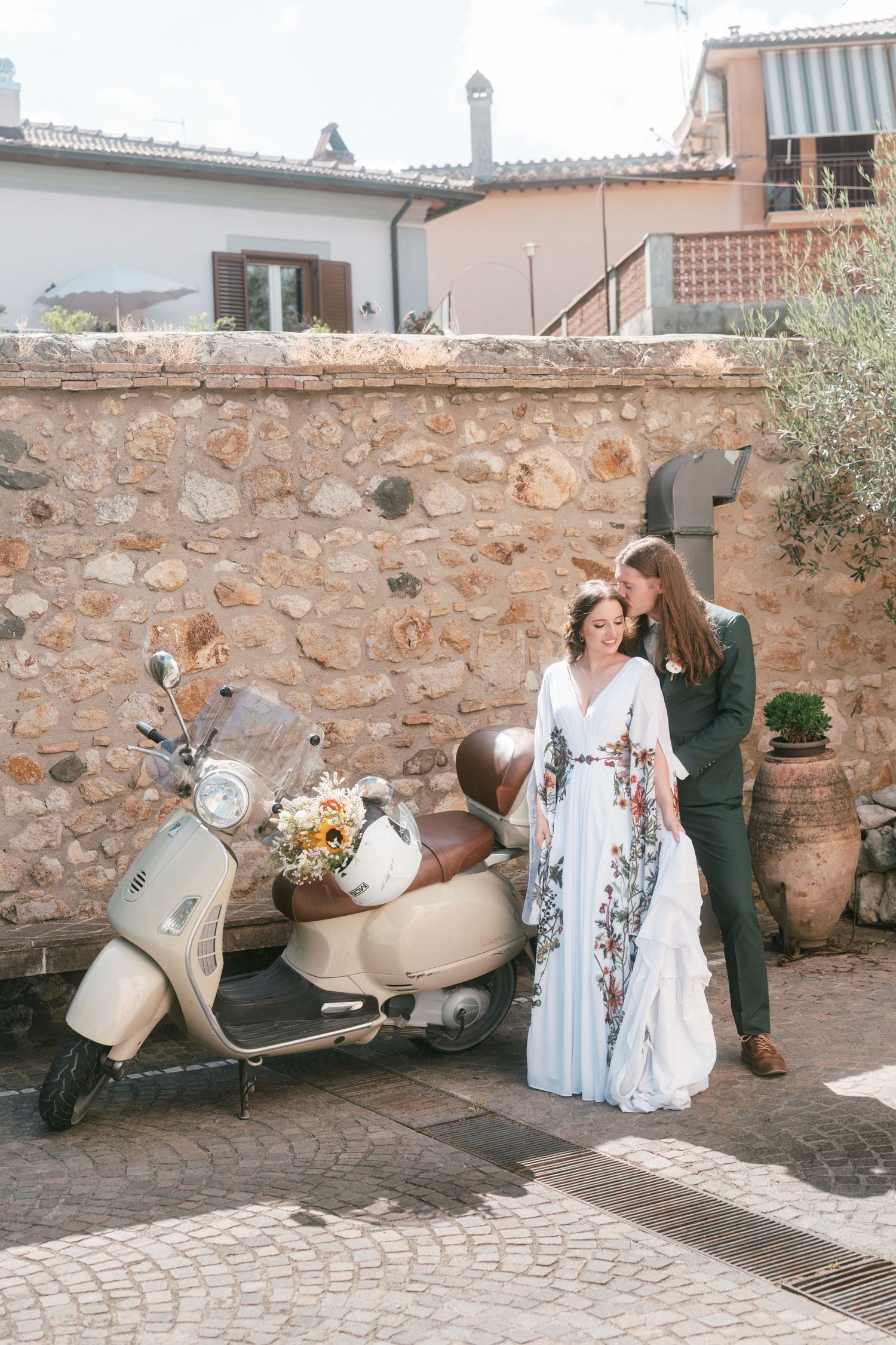 Couple in wedding attire posing beside a vintage scooter against a stone wall. Tuscany wedding photographer, filmmaker