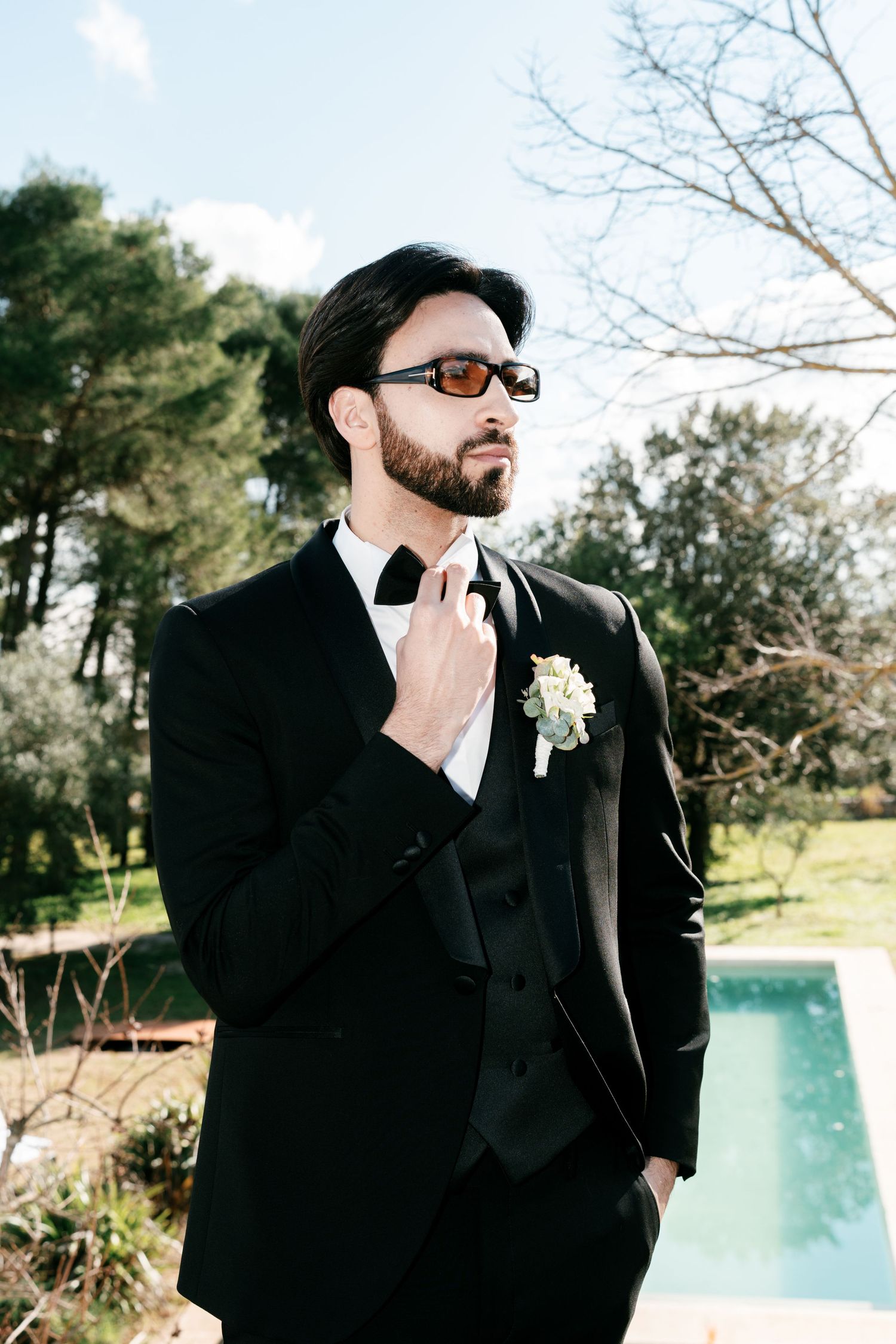 Dapper groom in black tuxedo with boutonniere and sunglasses standing near a pool in a garden. Puglia wedding photographer, film