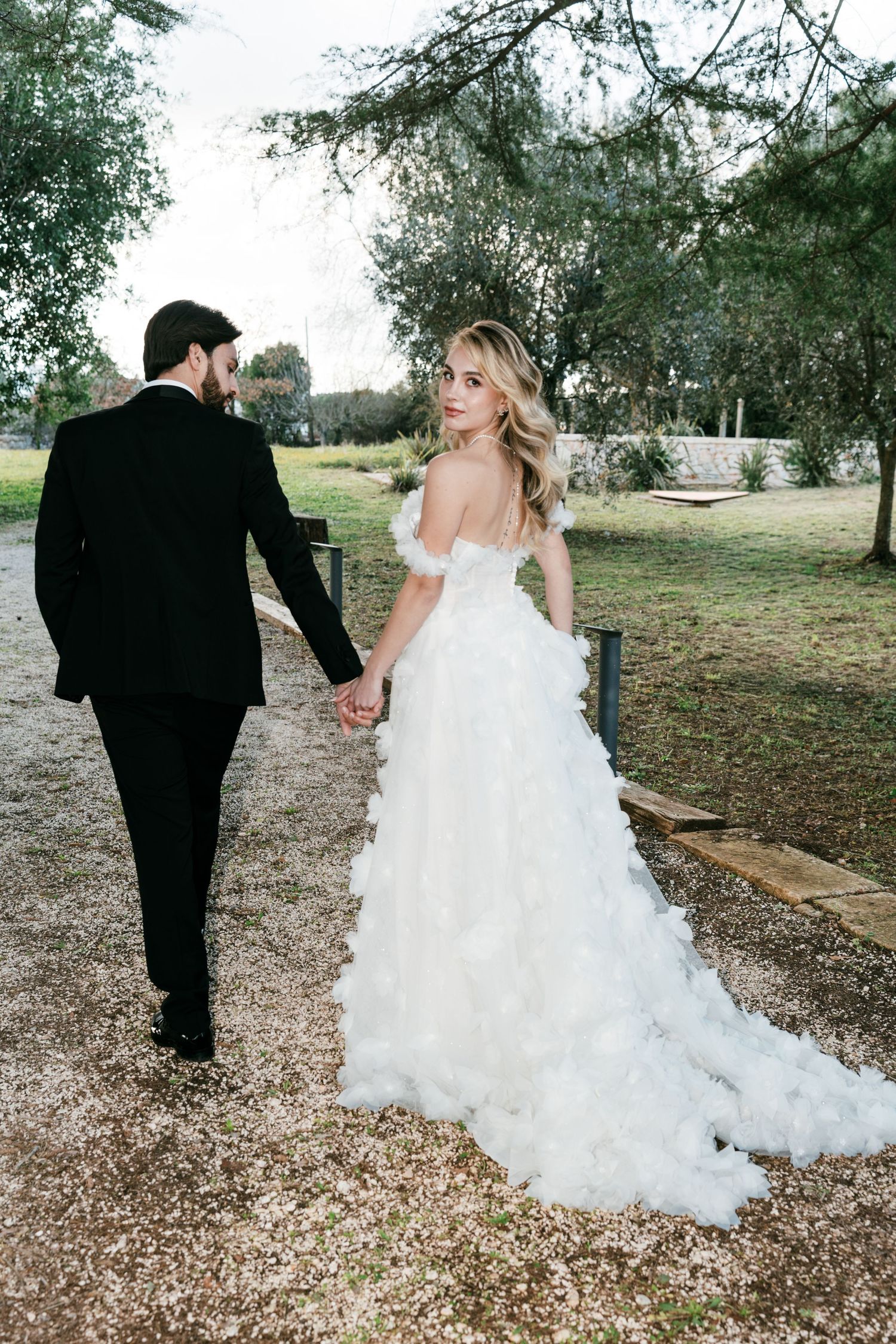 Bride in white ruffled off-shoulder gown holding hands with groom in black suit on garden path. Puglia wedding photographer, film