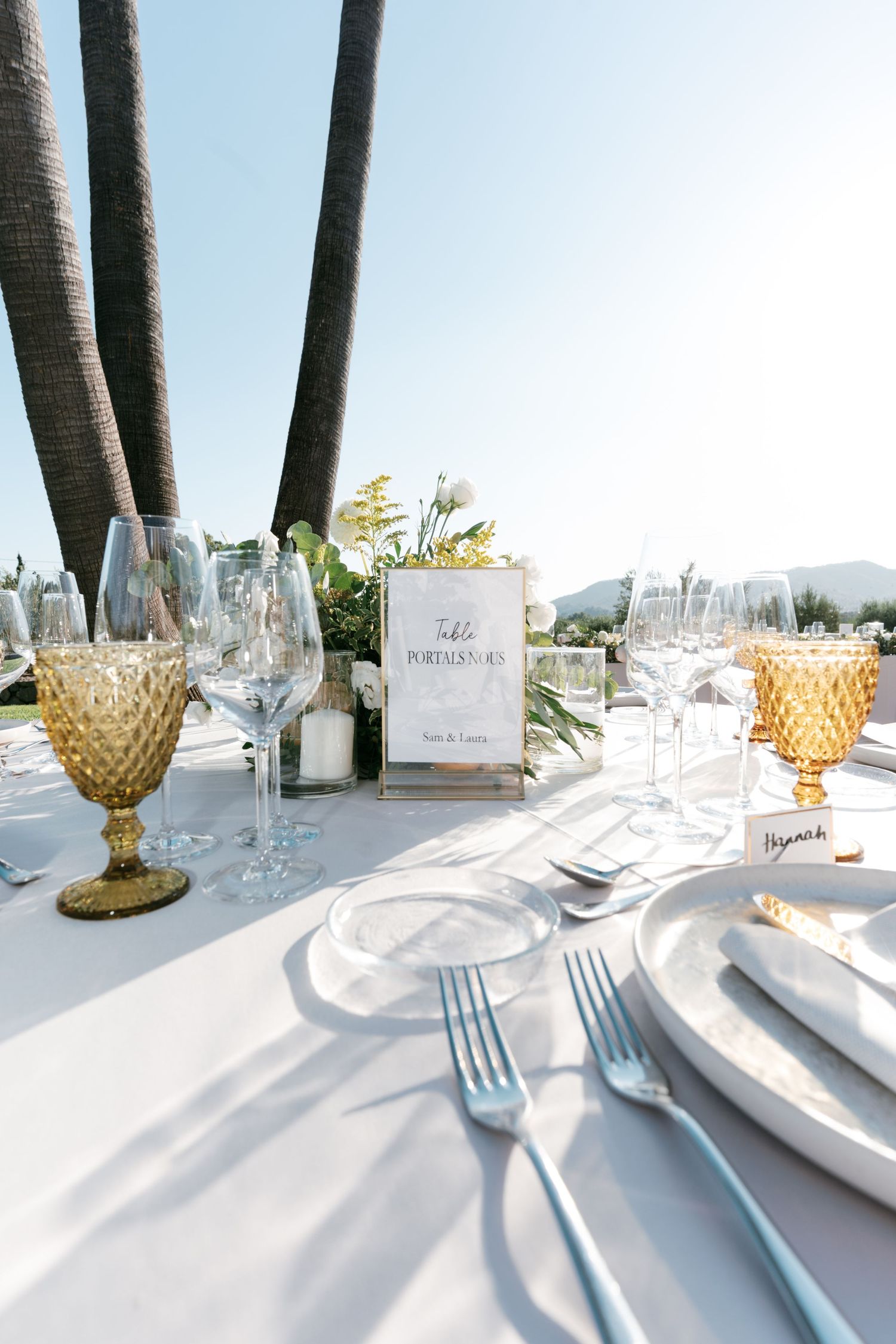 Elegant outdoor wedding table setting with amber goblets, white plates, and floral centerpiece under palm trees. Mallorca wedding photograph