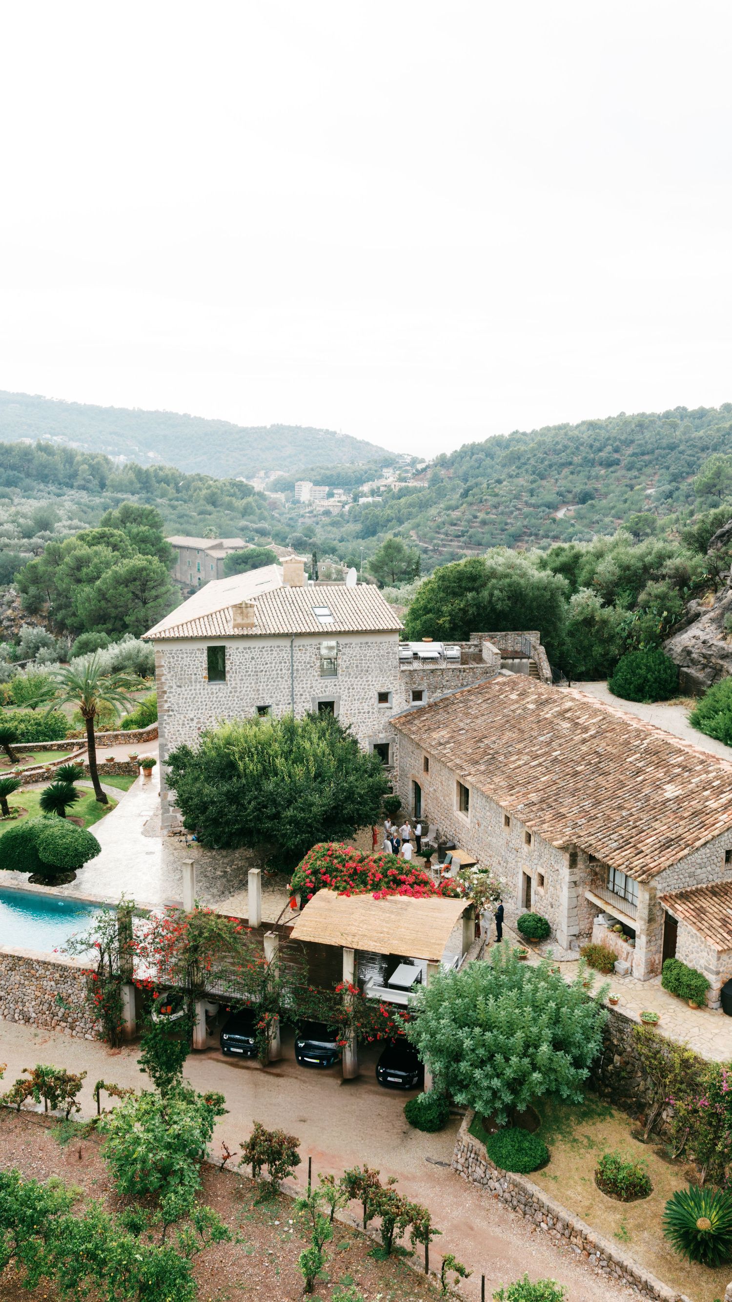 Aerial view of a rustic stone villa with terracotta roofs, swimming pool and lush gardens, set among forested Mediterranean hills.