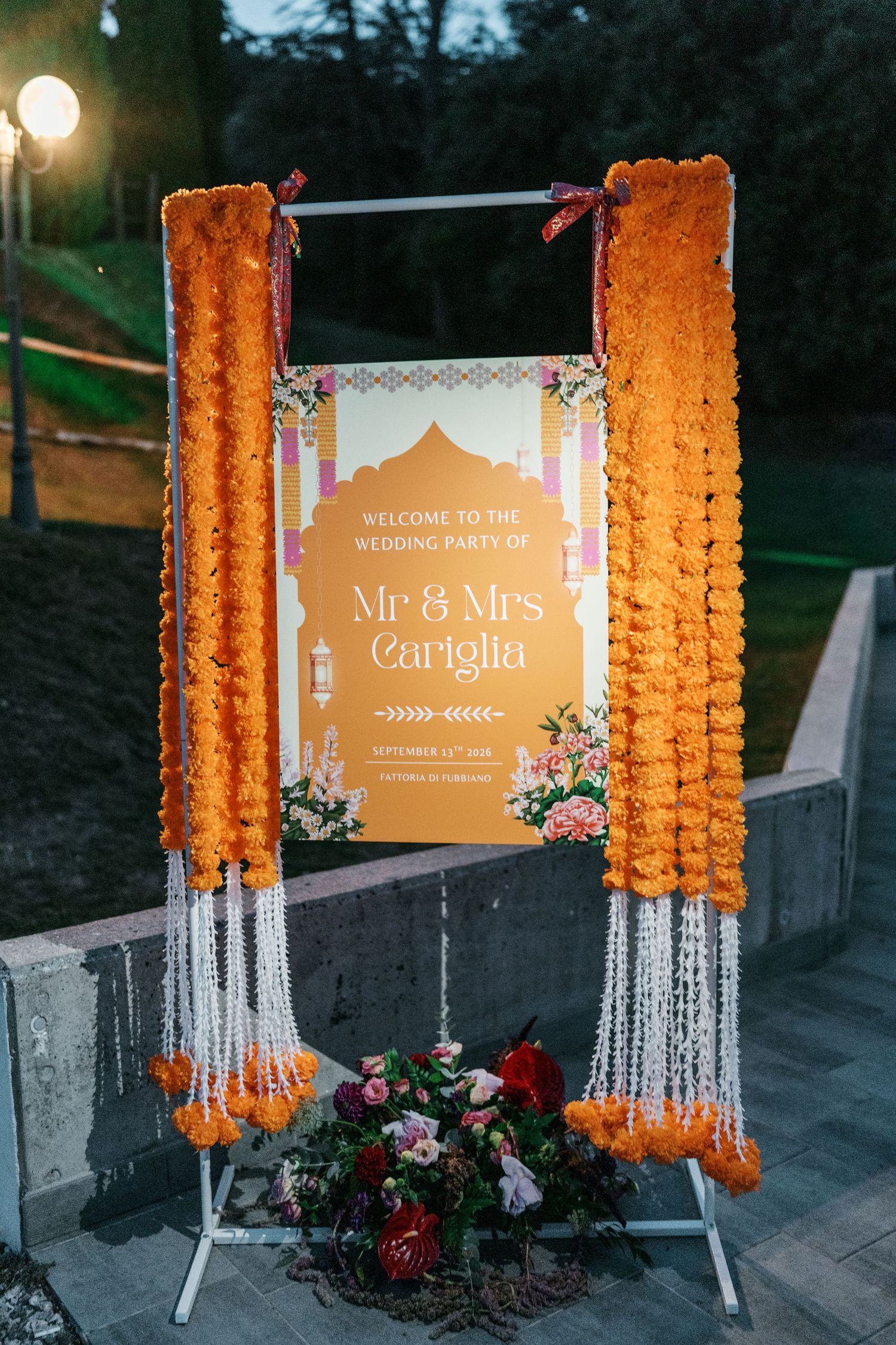 Wedding welcome sign for Mr & Mrs Cariglia framed by orange marigold garlands and a floral arrangement at dusk.