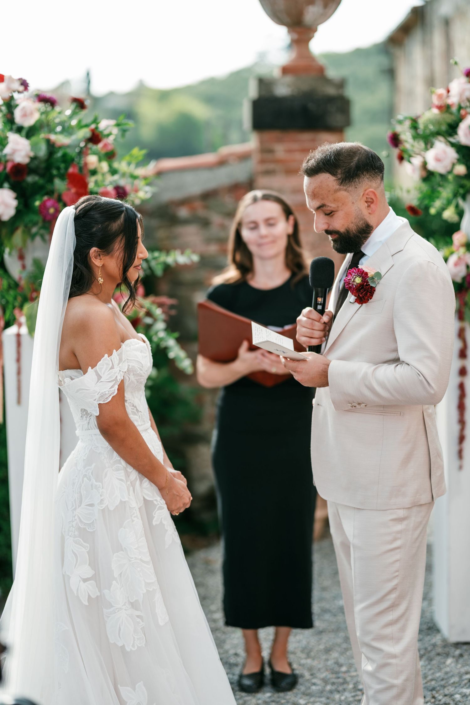 Groom in a cream suit reads vows into a microphone to his bride in a lace off-shoulder gown during an outdoor wedding ceremony.