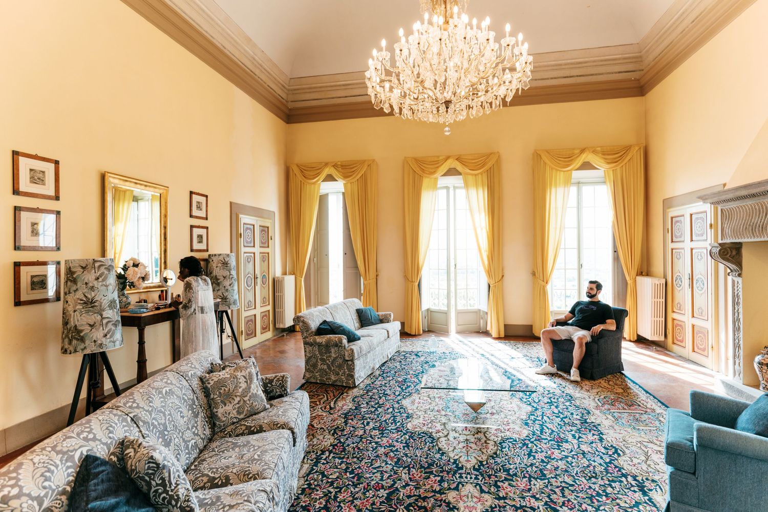 Elegant living room with a crystal chandelier, golden drapes, ornate patterned rug, and floral sofas, with a man seated by tall windows.