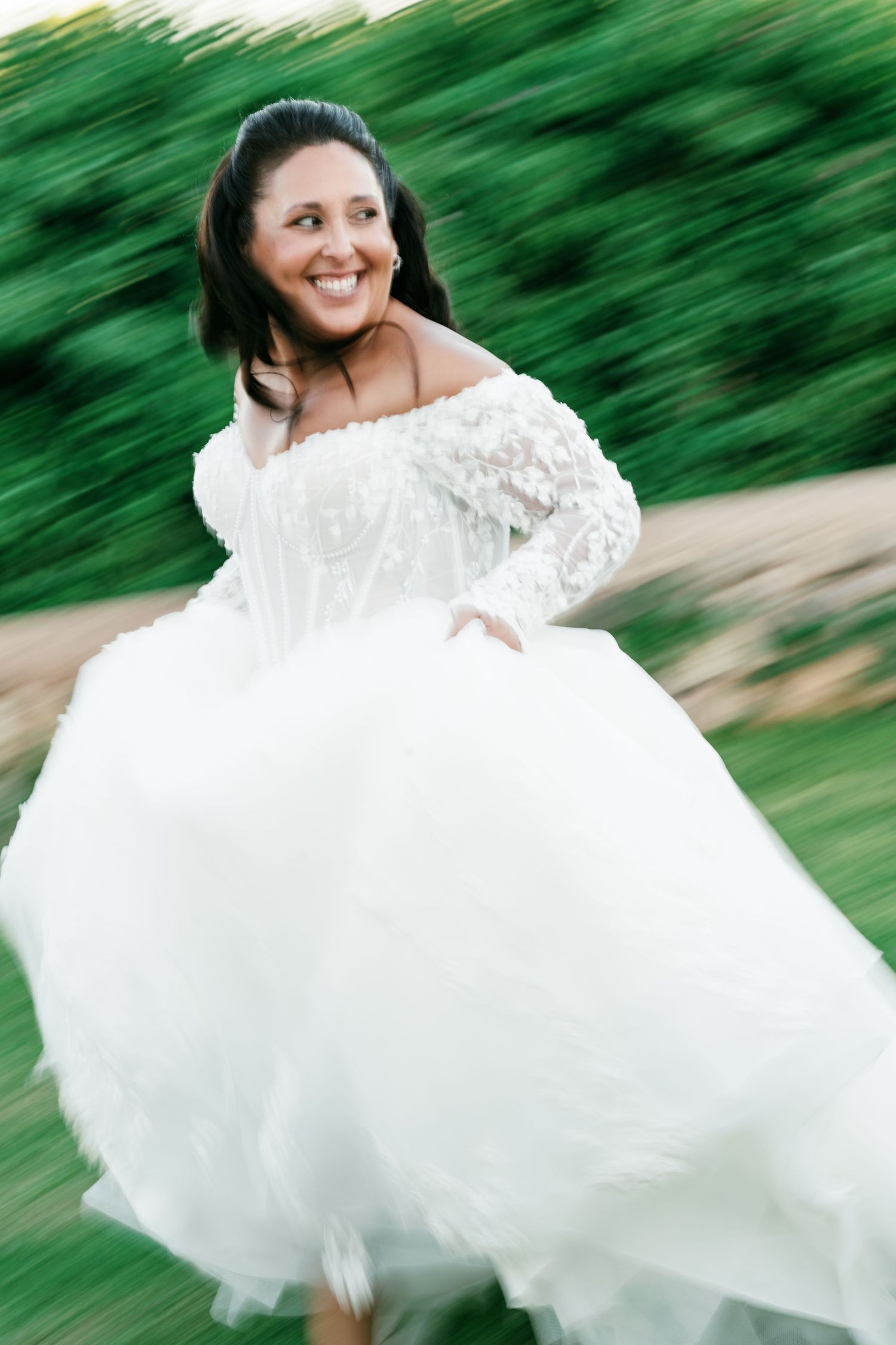 Smiling bride in an off-shoulder white lace wedding gown twirls outdoors on green grass, with a motion-blurred background.
