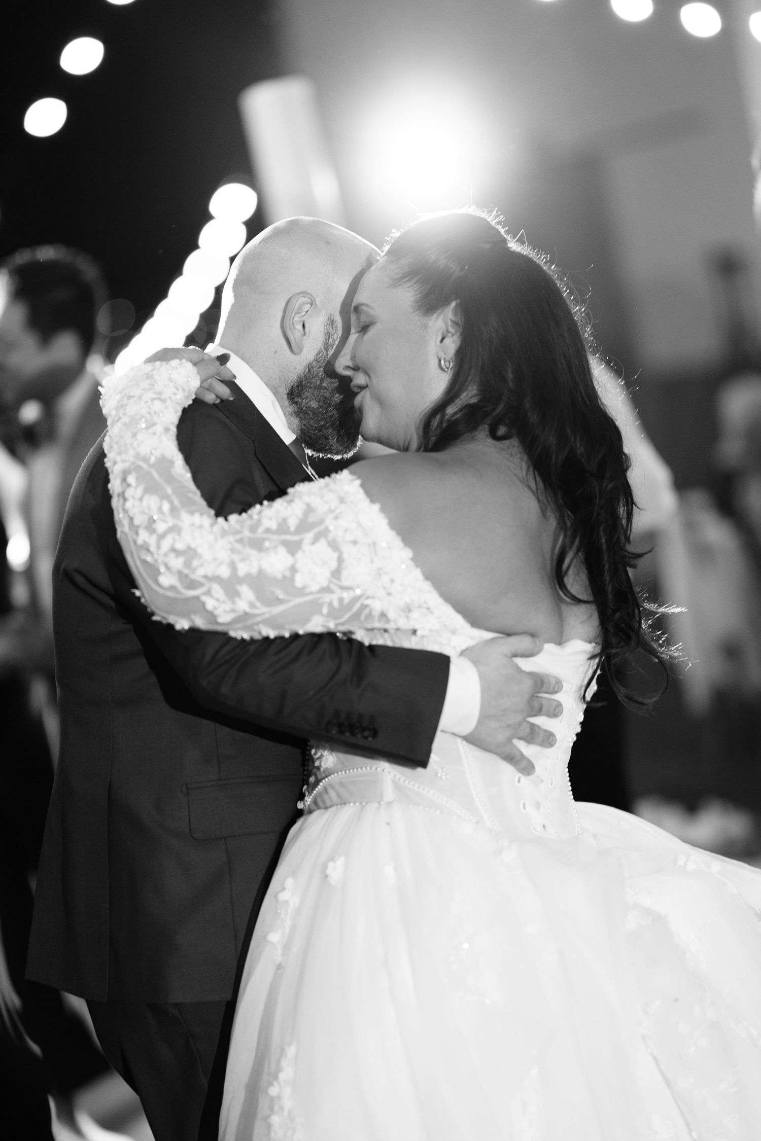 Black and white photo of a bride in a lace wedding gown and a groom in a suit sharing a close first dance under glowing string lights.