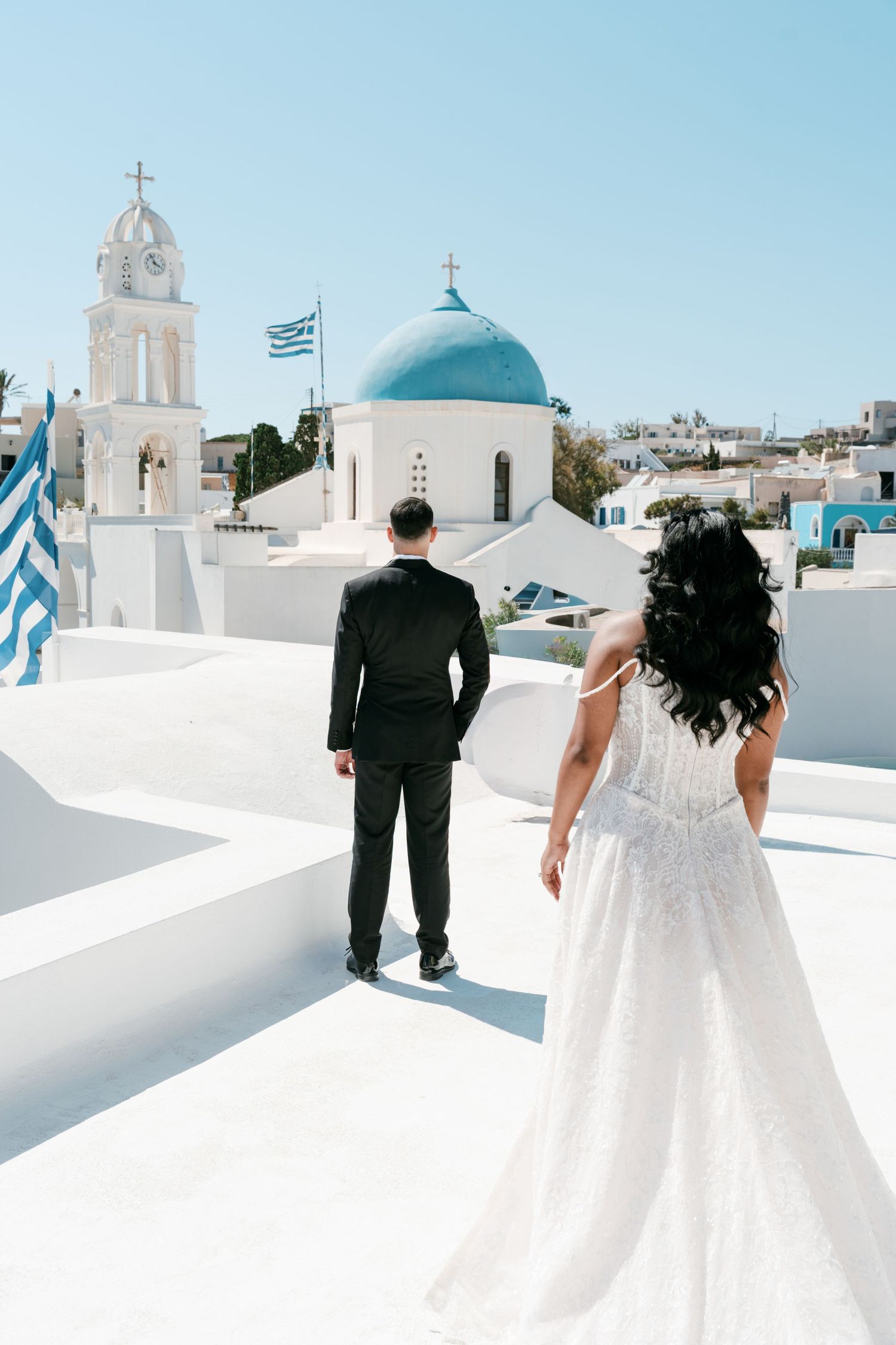 Bride and groom walk toward a white Greek Orthodox church with blue dome and Greek flag in sunny Santorini.