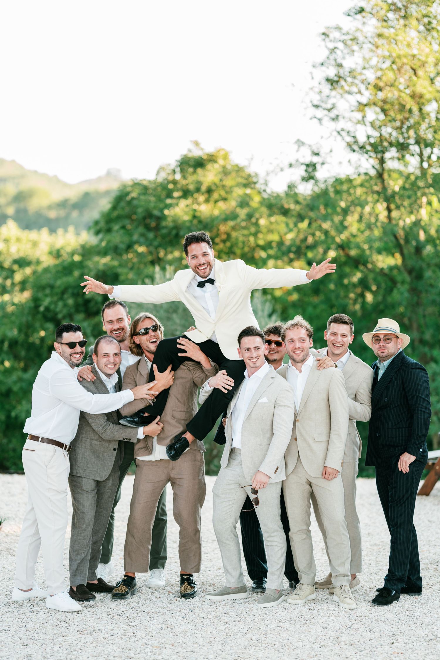 Groom in white tuxedo lifted by groomsmen in beige suits outdoors at a wedding celebration.