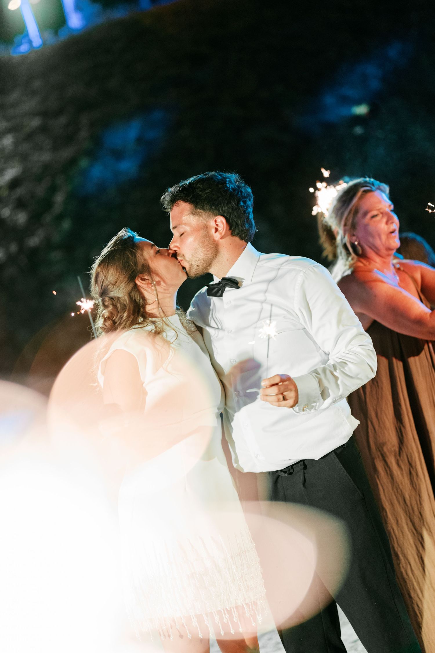 Newlyweds share a romantic kiss surrounded by sparklers at their outdoor evening wedding reception.