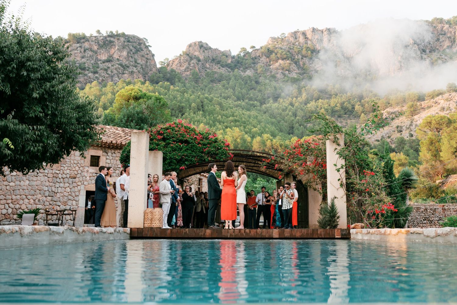 Wedding guests gather on a stone terrace beside a reflective pool, with misty forested mountains rising in the background.