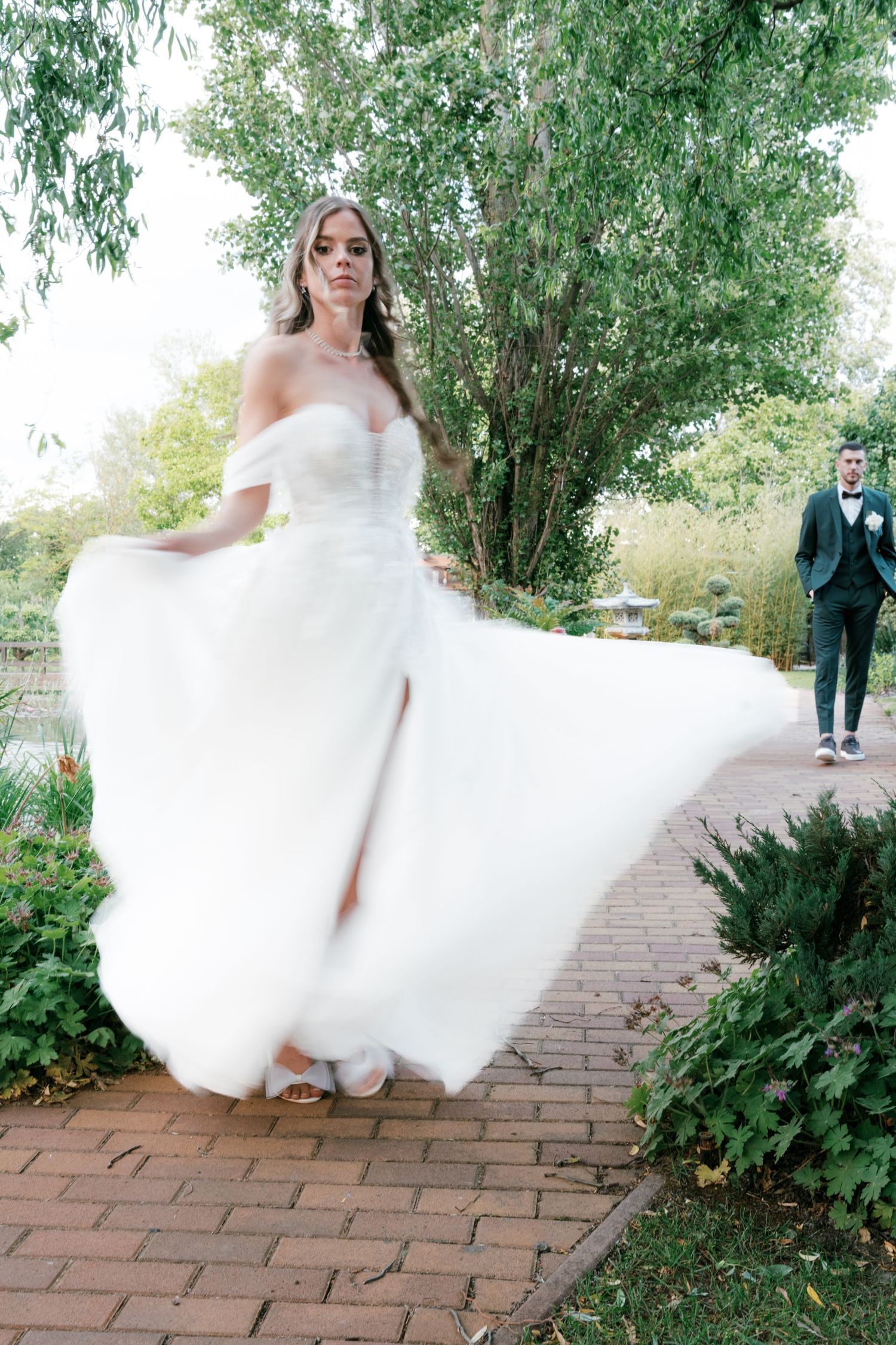Bride in off-shoulder white ball gown twirling on brick path with groom standing in background. Mallorca wedding photographer and filmmaker.