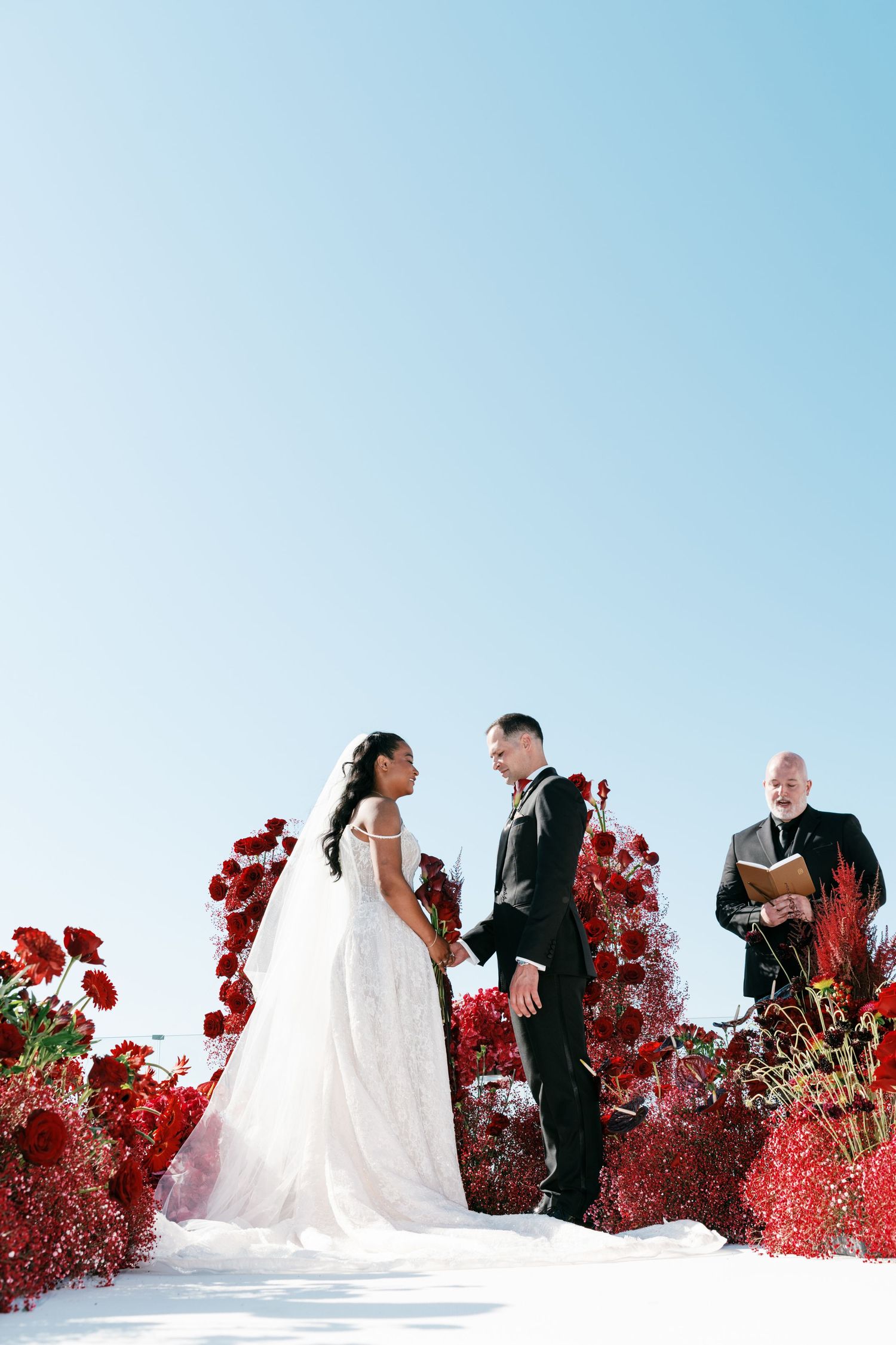 Bride and groom exchange vows outdoors surrounded by lush red floral arrangements under a clear blue sky.