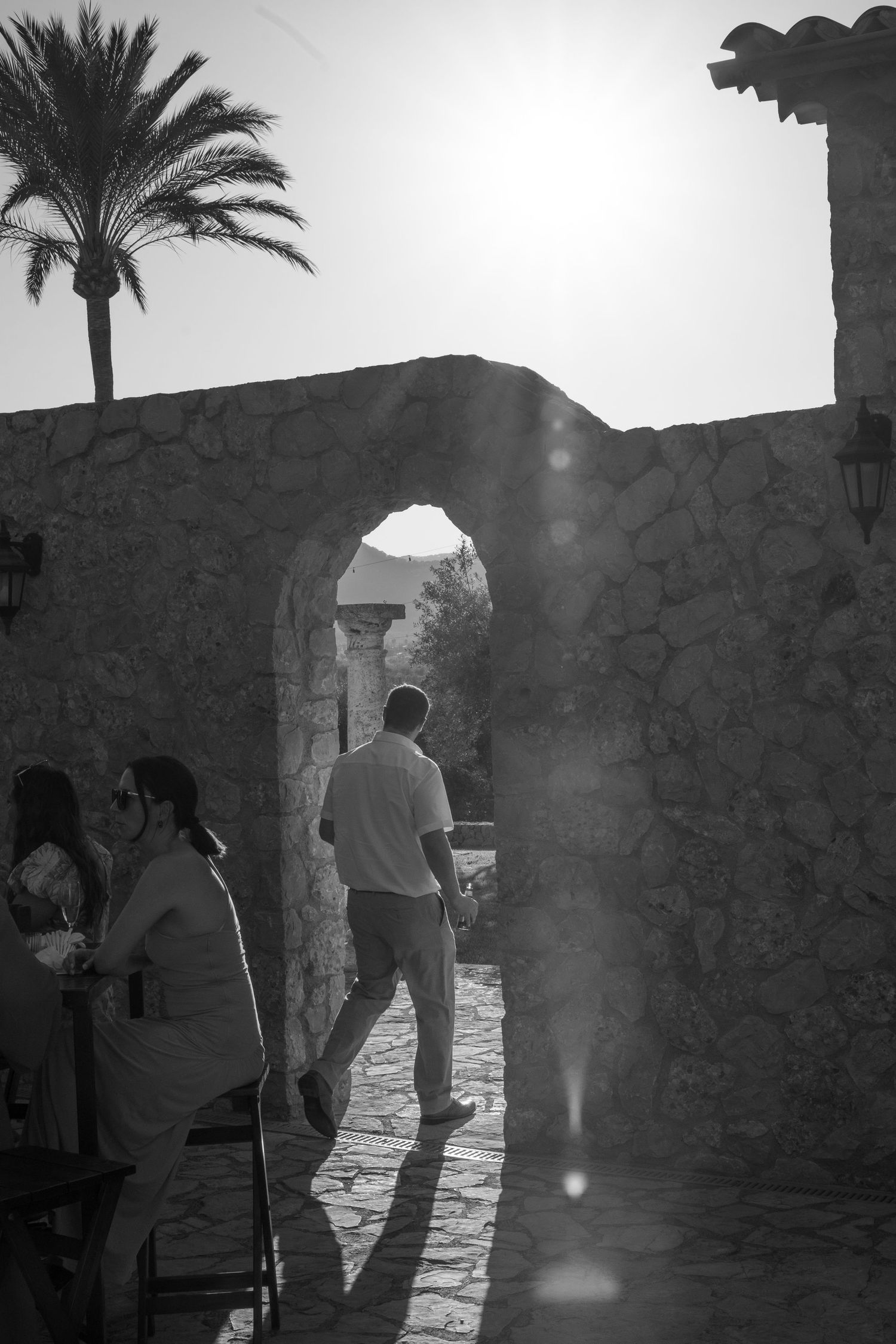 Black and white photo of a man walking through a stone archway with sun flare and palm tree silhouette. Mallorca wedding photographer, filmm