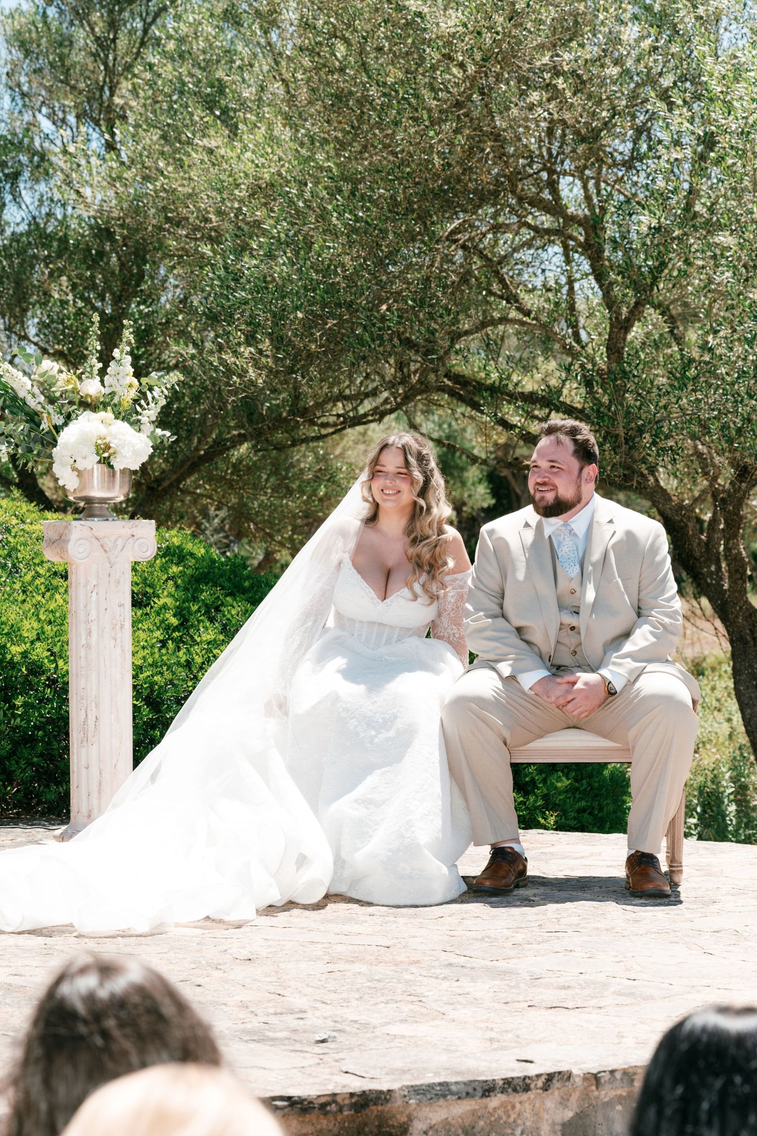 Bride in white gown and groom in tan suit sit together during outdoor wedding ceremony surrounded by olive trees in Mallorca.