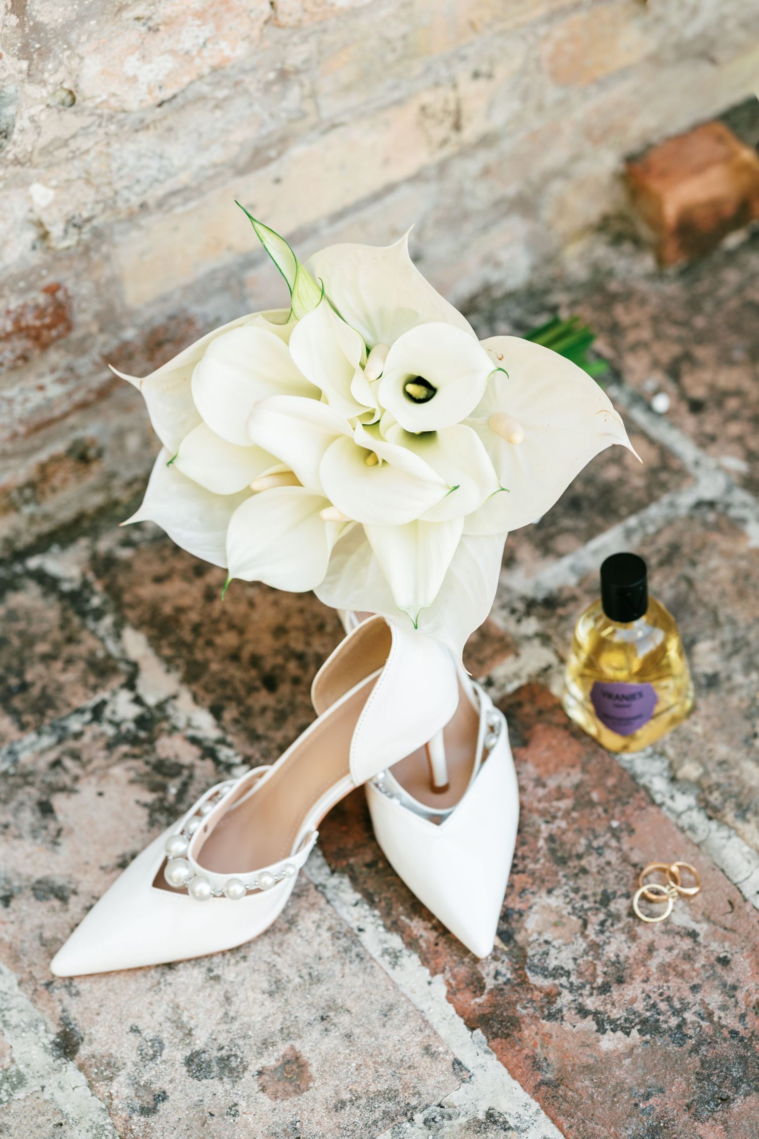 White bridal heels with pearl details beside white calla lily bouquet, gold rings, and perfume bottle.
