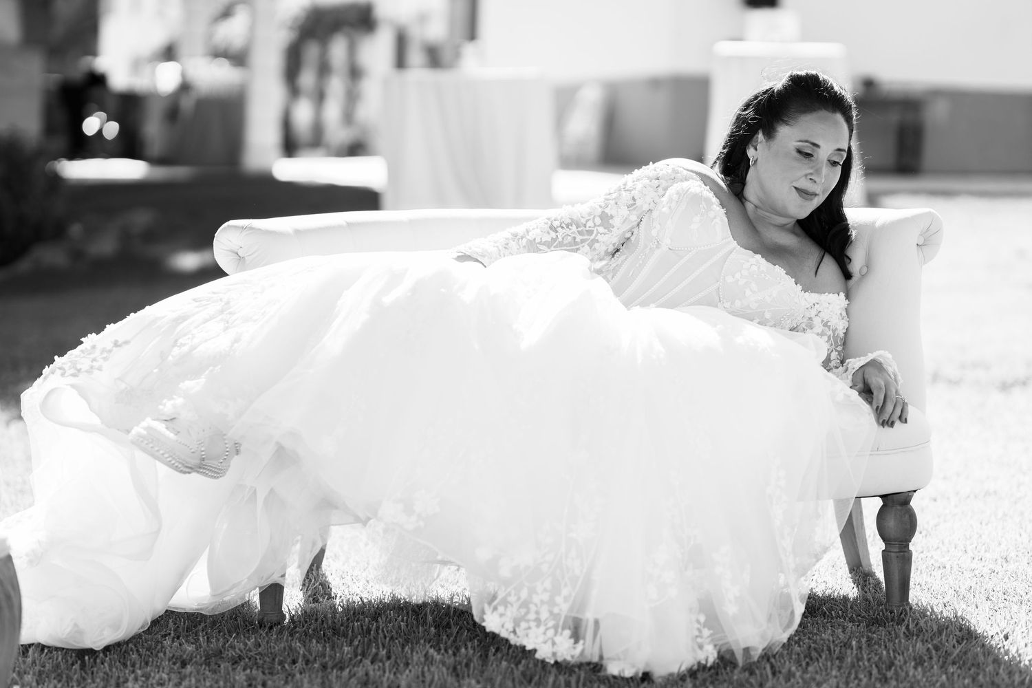 Black and white photo of a bride in a lace wedding gown reclining on a white vintage sofa placed on a grassy lawn.