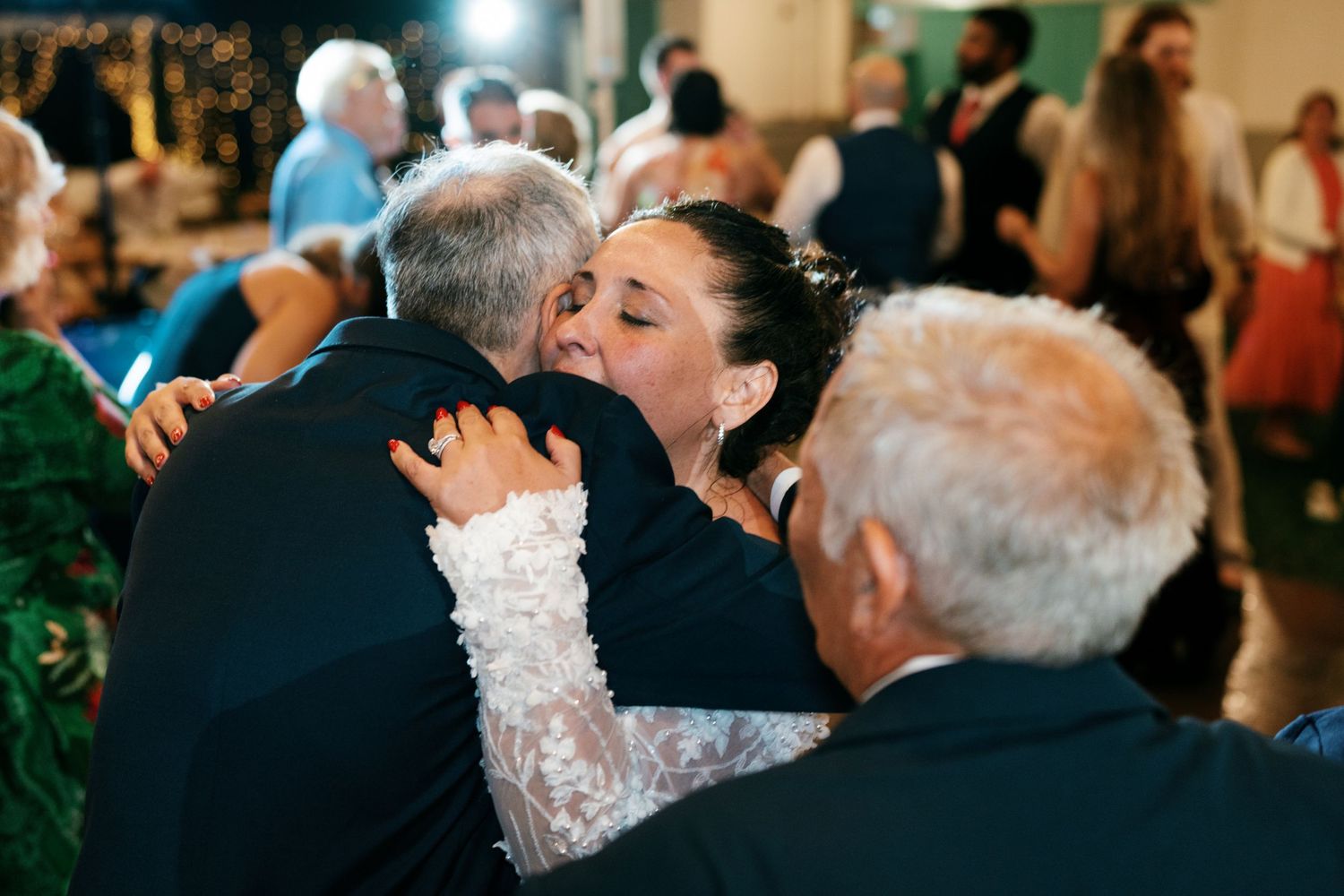 Bride in a white lace gown shares an emotional embrace with an older man during a wedding reception as guests look on.