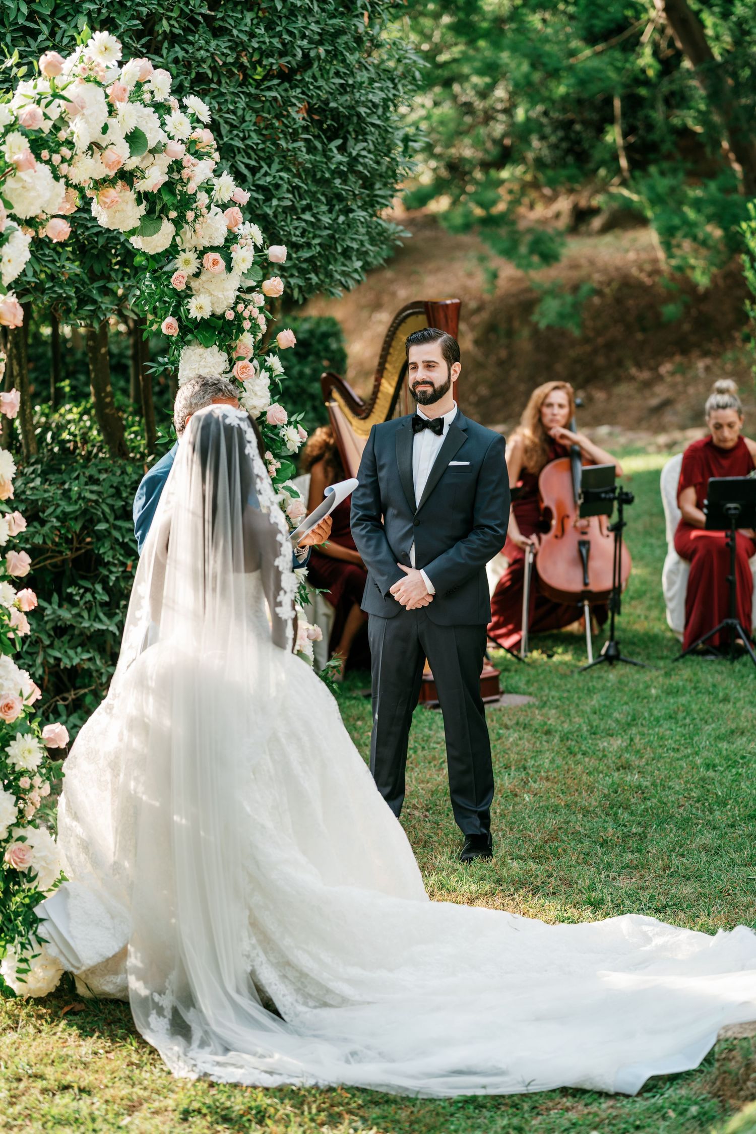 Bride in a long-veil wedding gown faces a groom in a tuxedo during an outdoor garden ceremony with floral arch and string musicians.