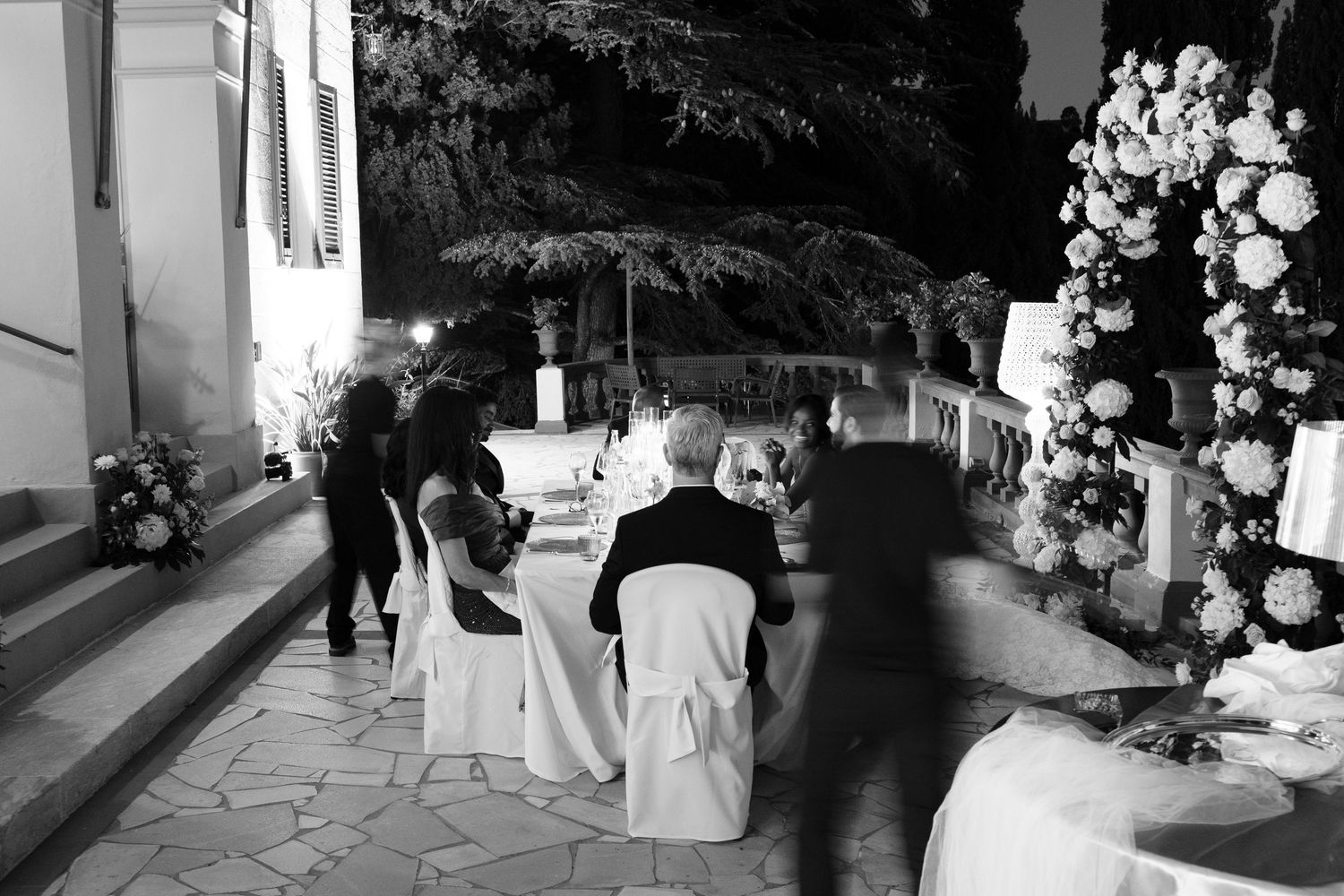 Black and white scene of guests seated at a candlelit outdoor wedding dinner table beside floral arrangements at night.