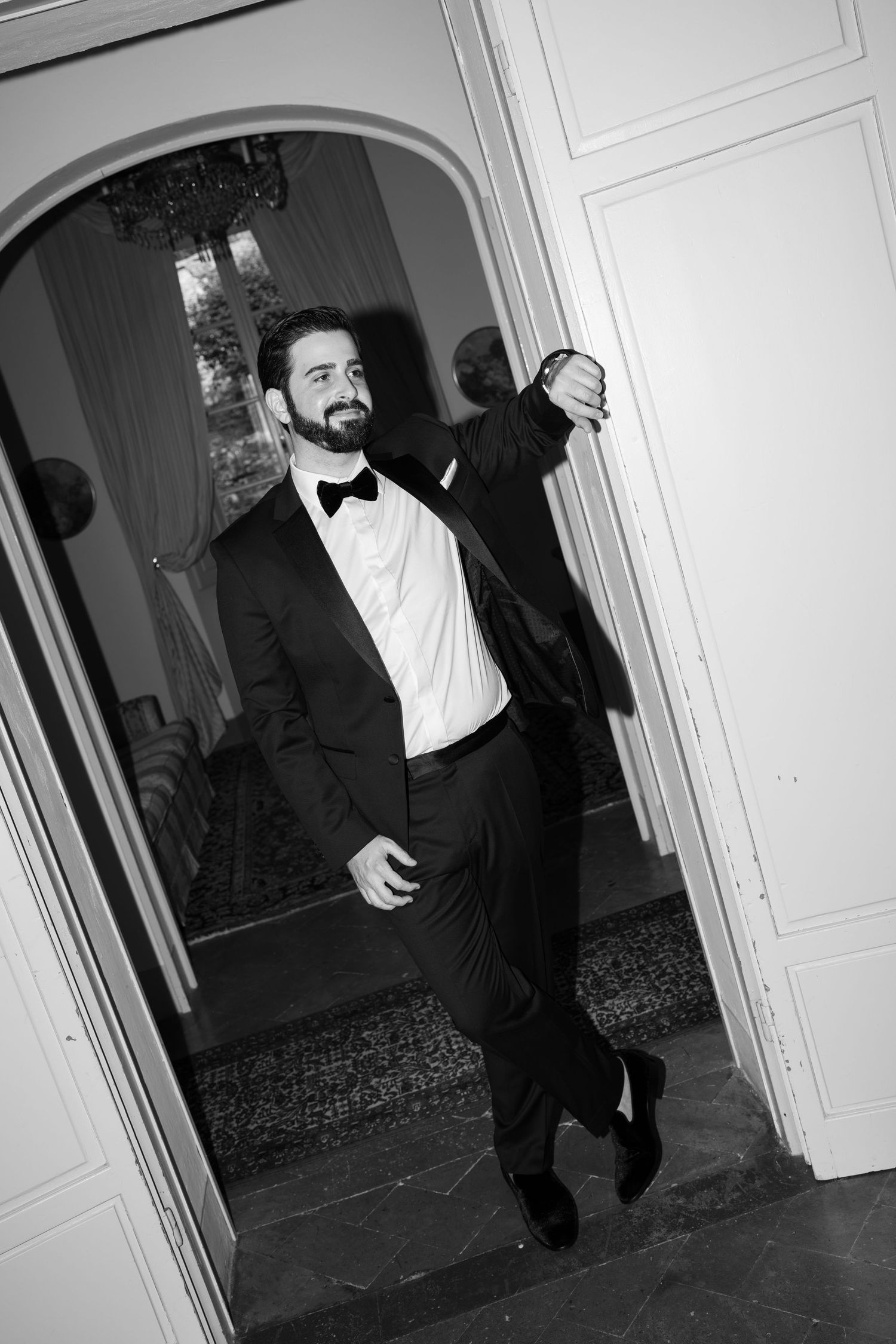 Bearded man in a black tuxedo and bow tie leans casually against an arched doorway in a black and white formal portrait.