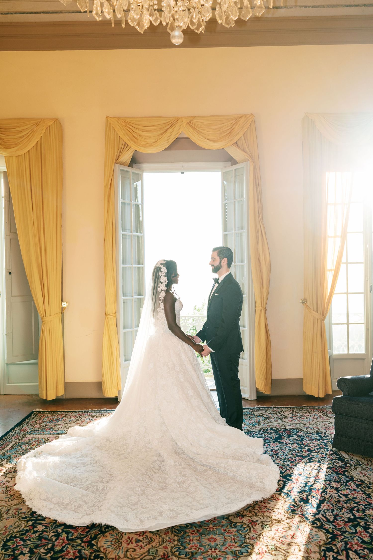 Bride in a lace gown with a long train holds hands with her groom in a tuxedo before sunlit windows in an elegant wedding venue.