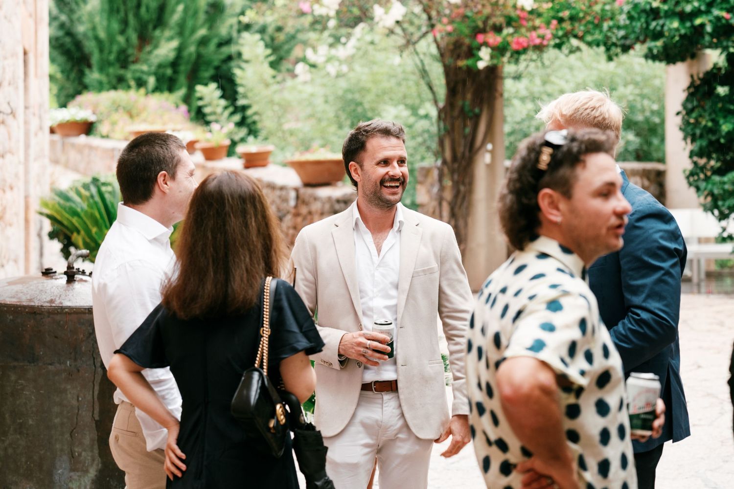 Smiling man in a beige blazer chats with friends at an outdoor garden party, surrounded by greenery and stone walls.