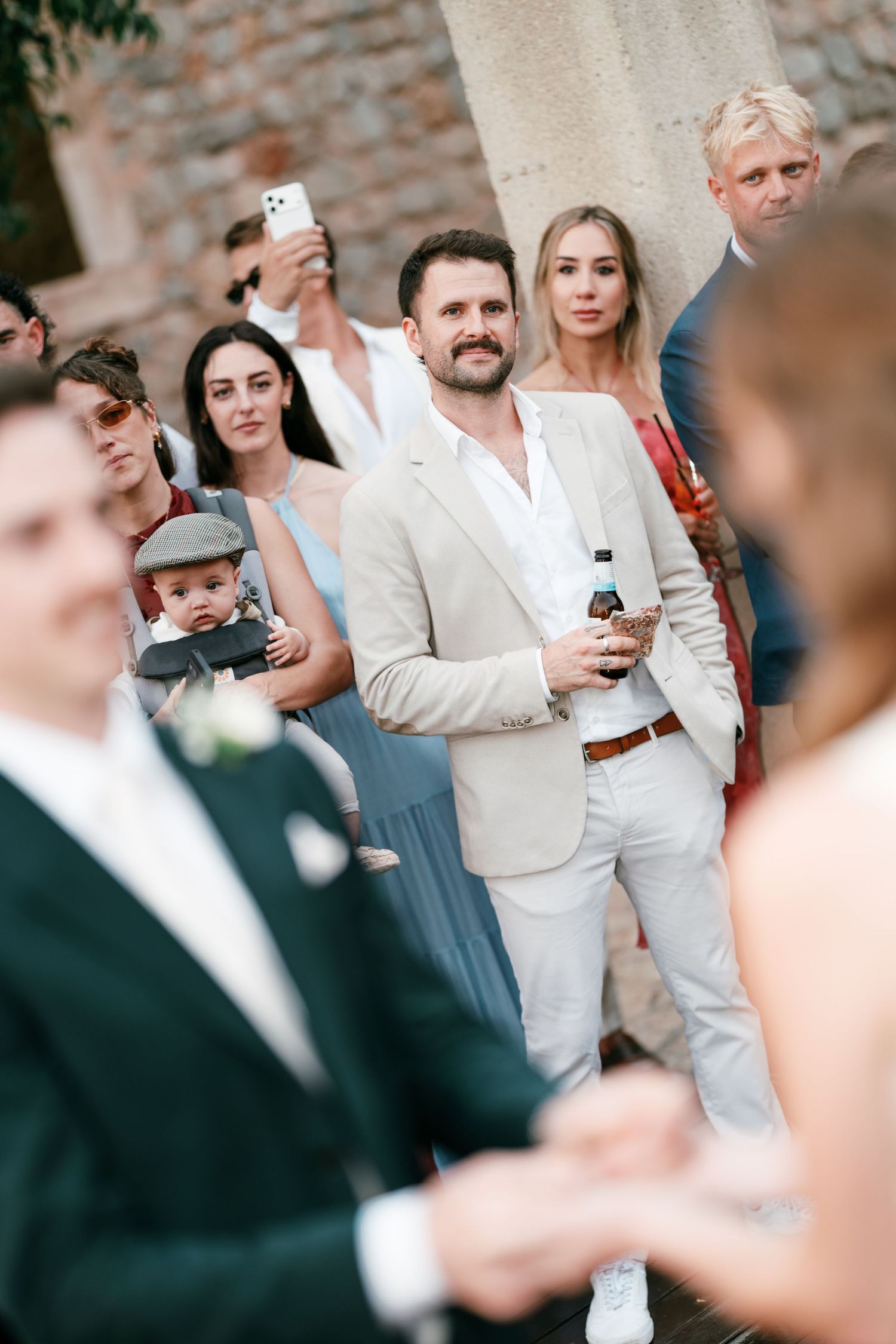 Smiling bearded guest in a beige suit holds a drink while watching a wedding ceremony alongside other attendees.