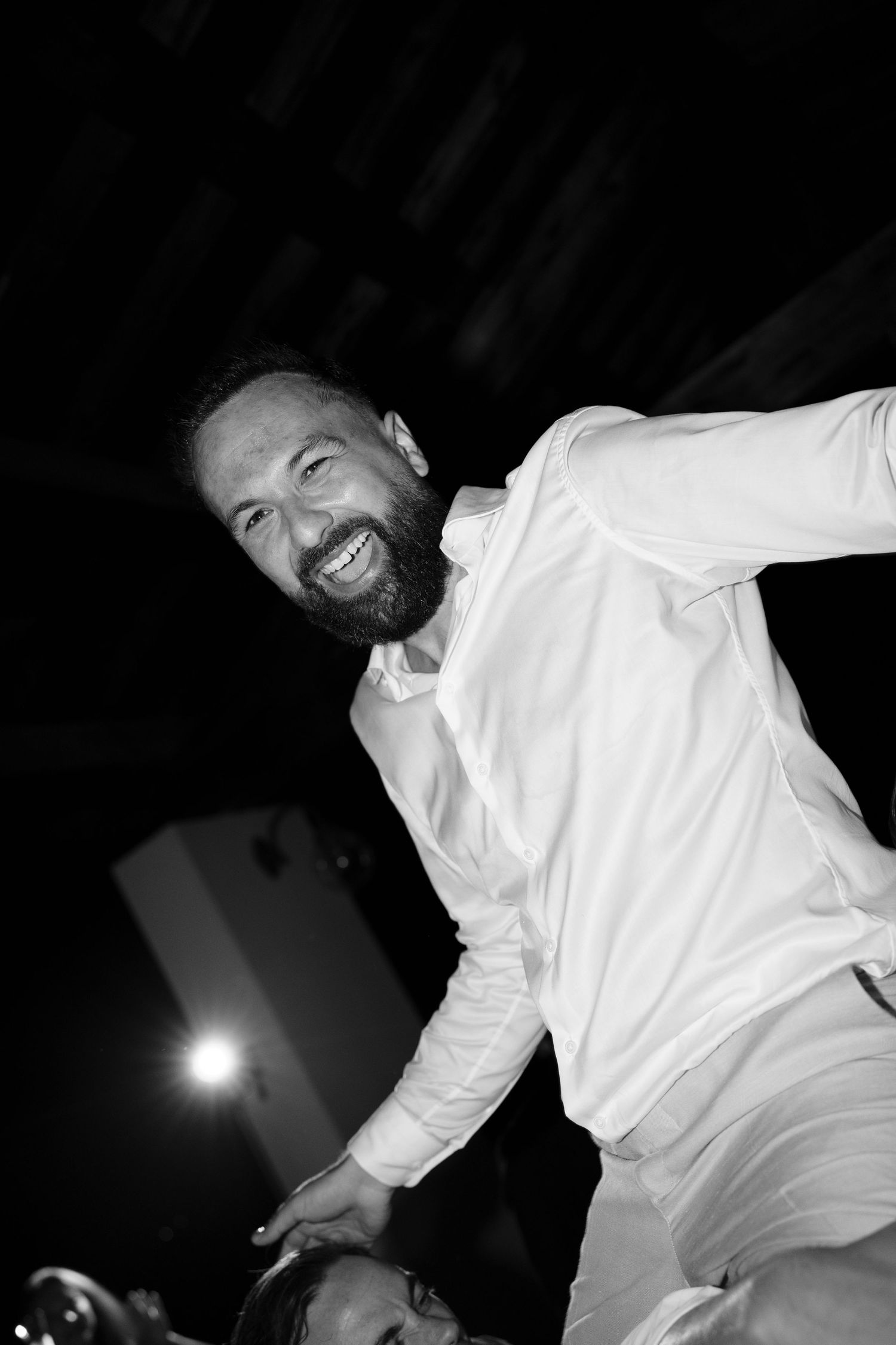 Black and white photo of a smiling bearded man in a white shirt leaning forward and laughing at a lively evening celebration.
