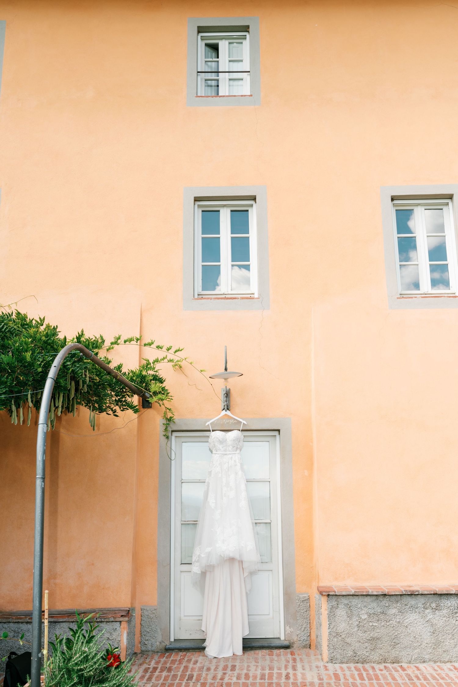 White lace wedding dress hanging on a vintage door of a peach-colored Tuscan villa with a green vine pergola and brick patio.
