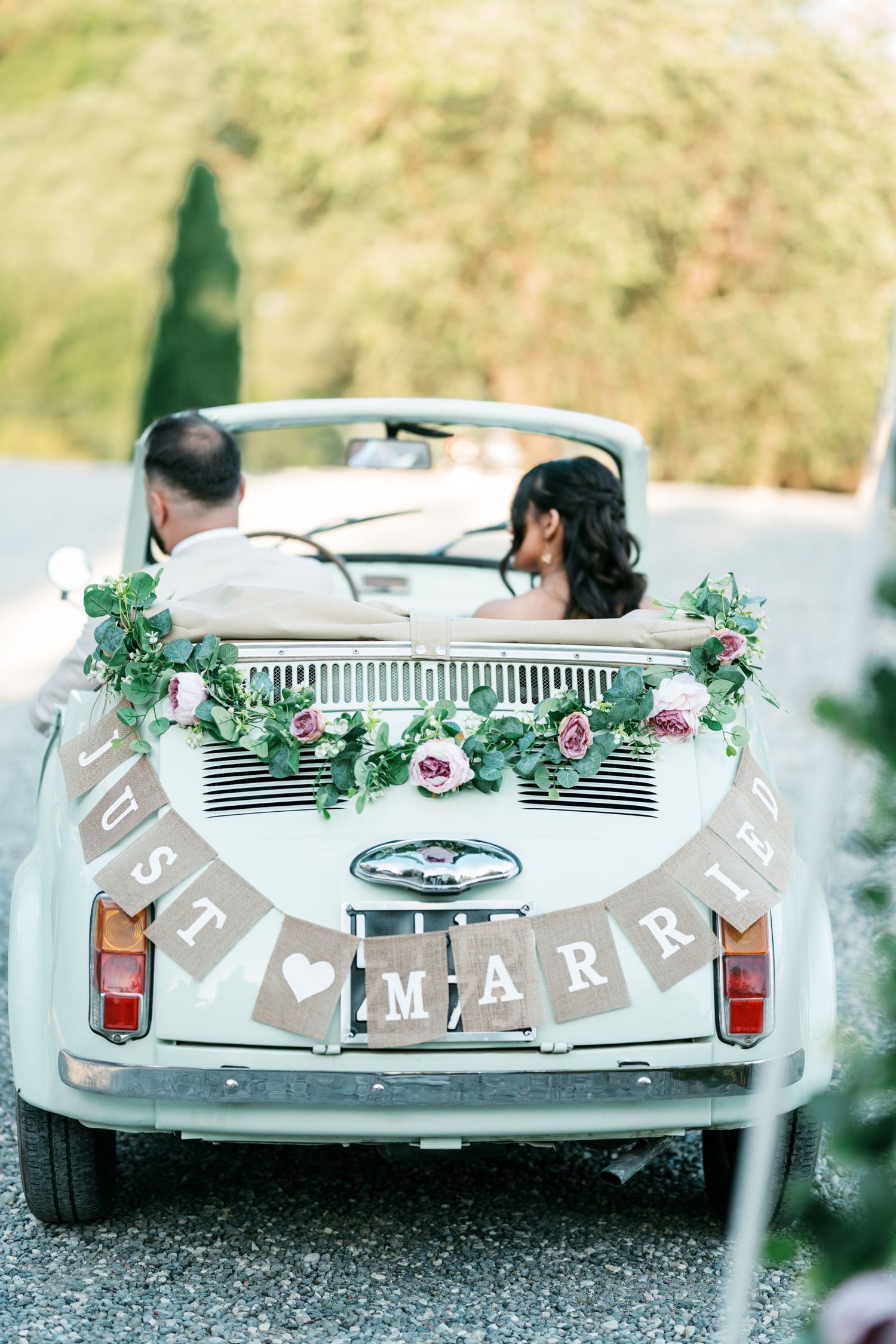 Vintage white convertible decorated with pink rose garland and a 'Just Married' burlap banner as a couple drives away on a gravel road.