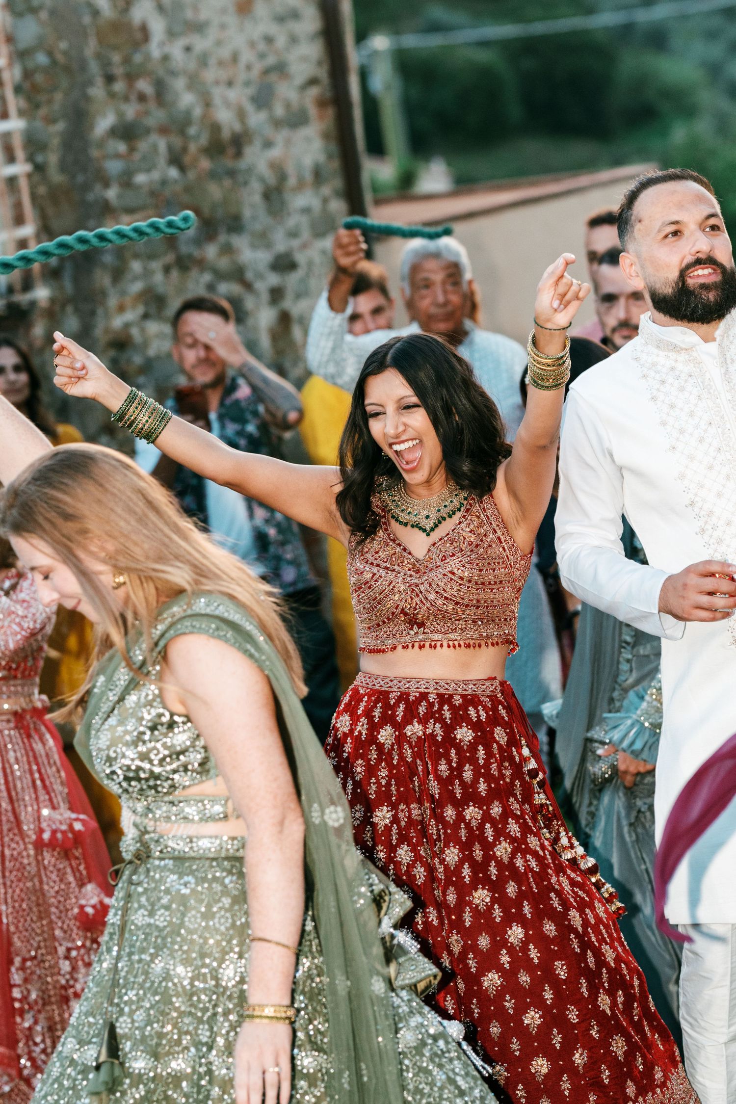 Laughing bride in a red and gold lehenga dances joyfully with arms raised as wedding guests celebrate around her.