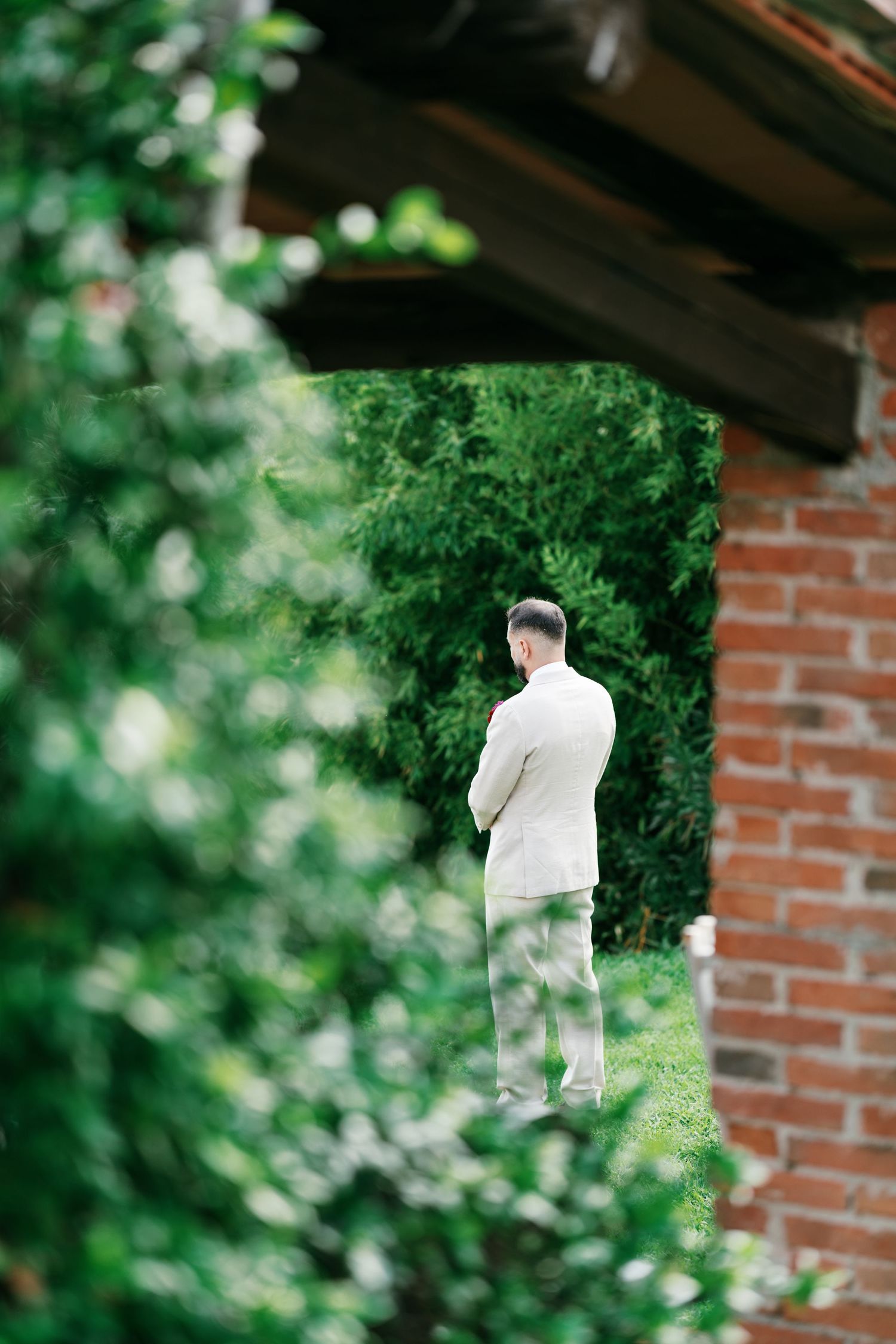 Man in a light beige suit stands with his back to the camera, framed by a brick archway and blooming white flowers in a green garden.