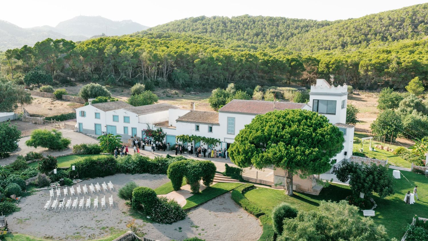 Aerial view of a white Mediterranean estate venue with terracotta roofs, manicured gardens, outdoor seating and pine-covered hills beyond.