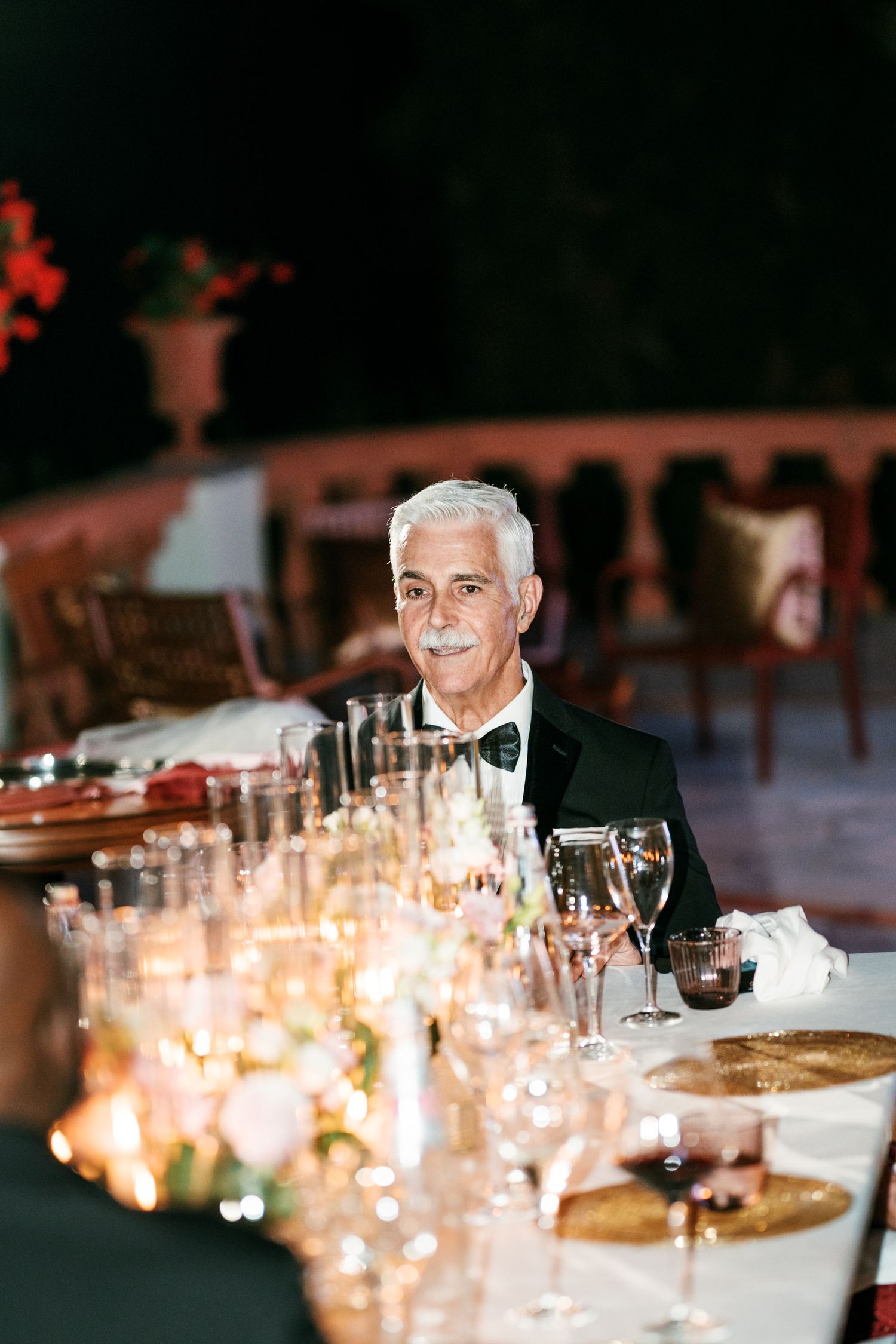 Elderly man with white hair, mustache and bow tie sits at a candlelit wedding reception table with glasses and floral decor.