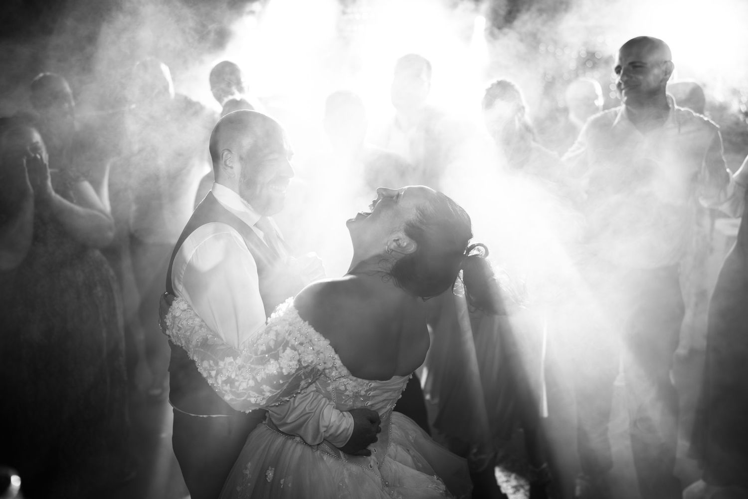 Black and white photo of a bride in an off-the-shoulder lace gown leaning back in a dance dip as backlit guests look on through haze.