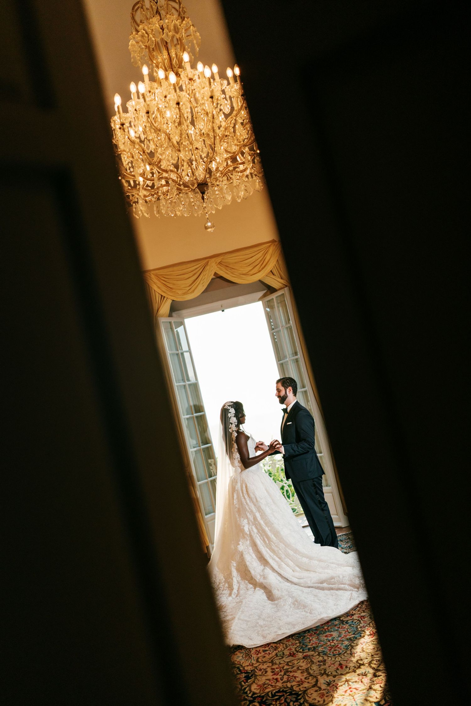 Bride in a flowing ball gown and groom in a dark suit stand holding hands by tall windows beneath a crystal chandelier.