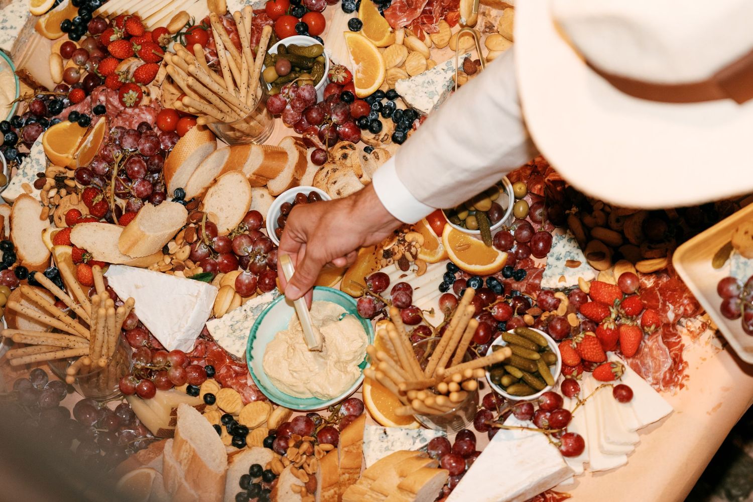 Person in a white hat reaches for a large charcuterie grazing table with grapes, tomatoes, citrus slices, breadsticks, crackers, and dips.