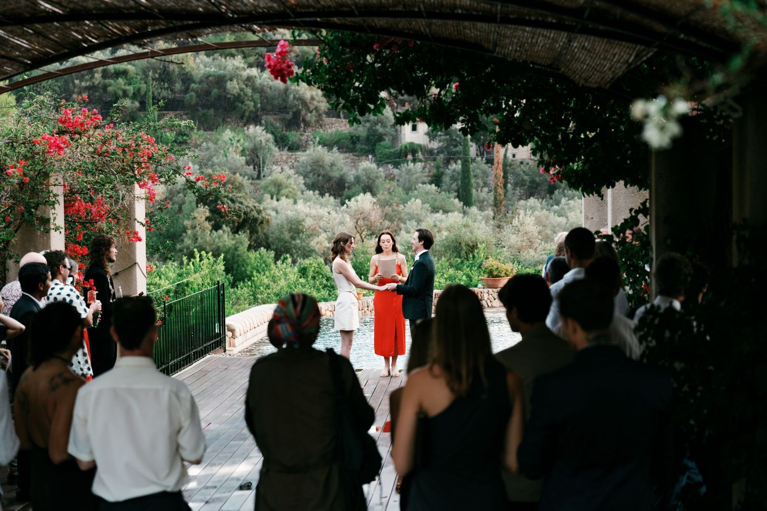 Wedding guests watch an outdoor ceremony as a bride, groom and officiant stand beneath a vine-covered pergola overlooking a lush garden.