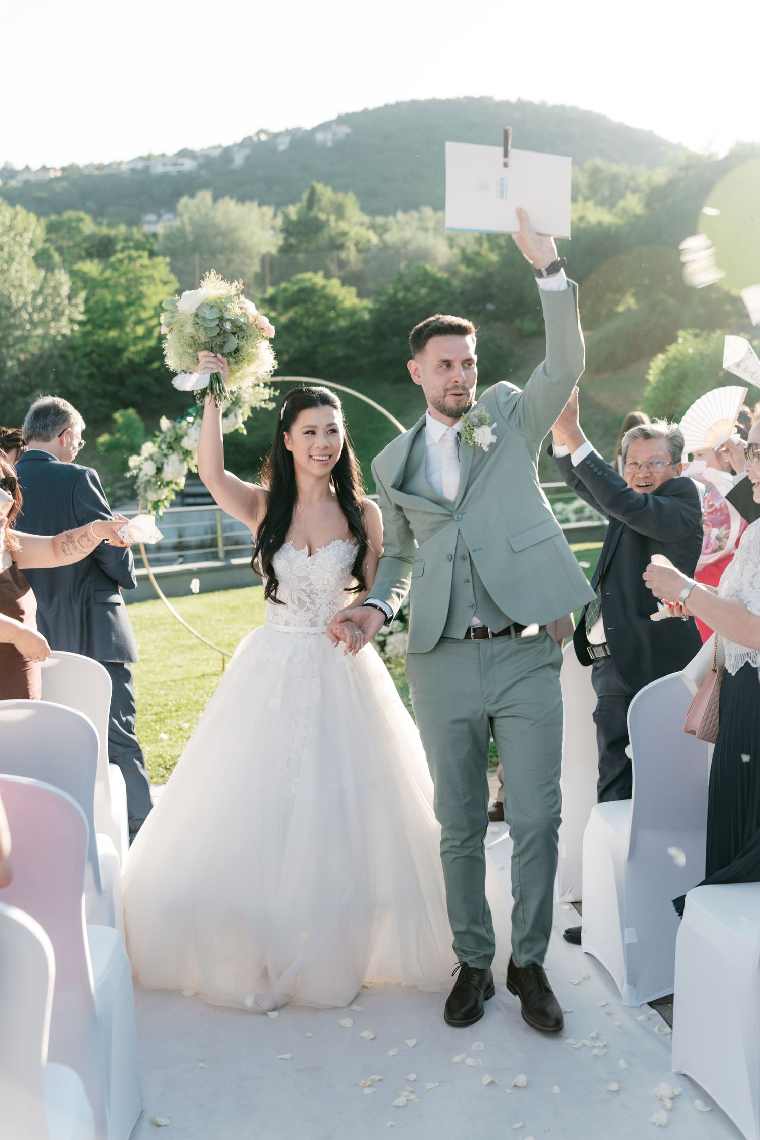Newlyweds celebrate their recessional, raising bouquet and marriage certificate while guests cheer outdoors. Italy wedding photographer.